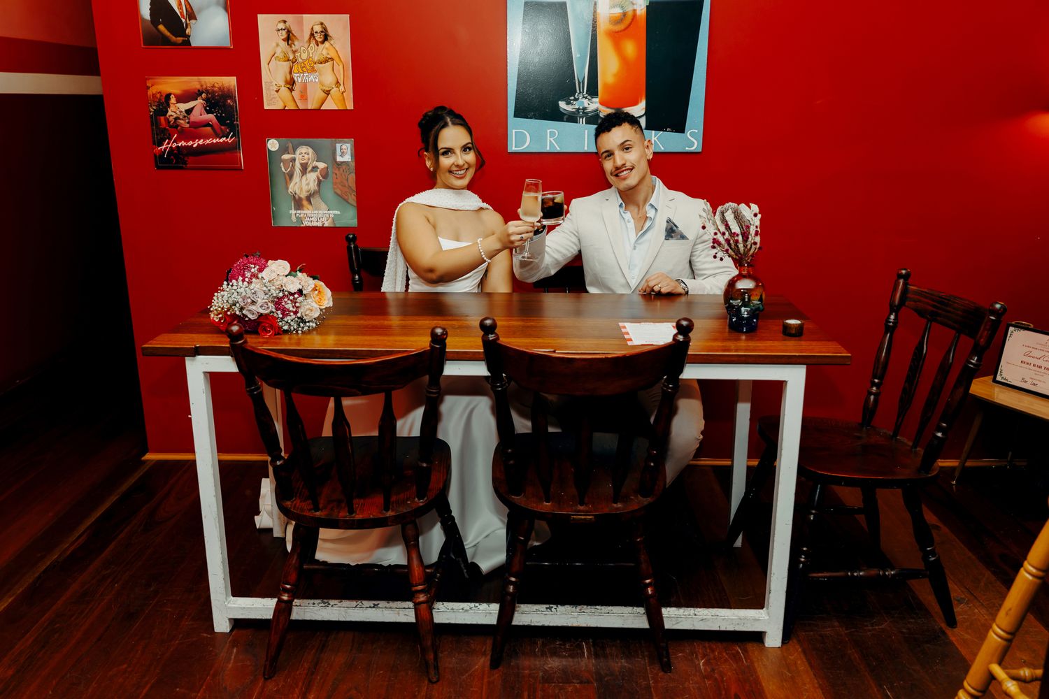Newlywed couple sitting at a wooden table in a red-walled restaurant, smiling and raising glasses in a toast.