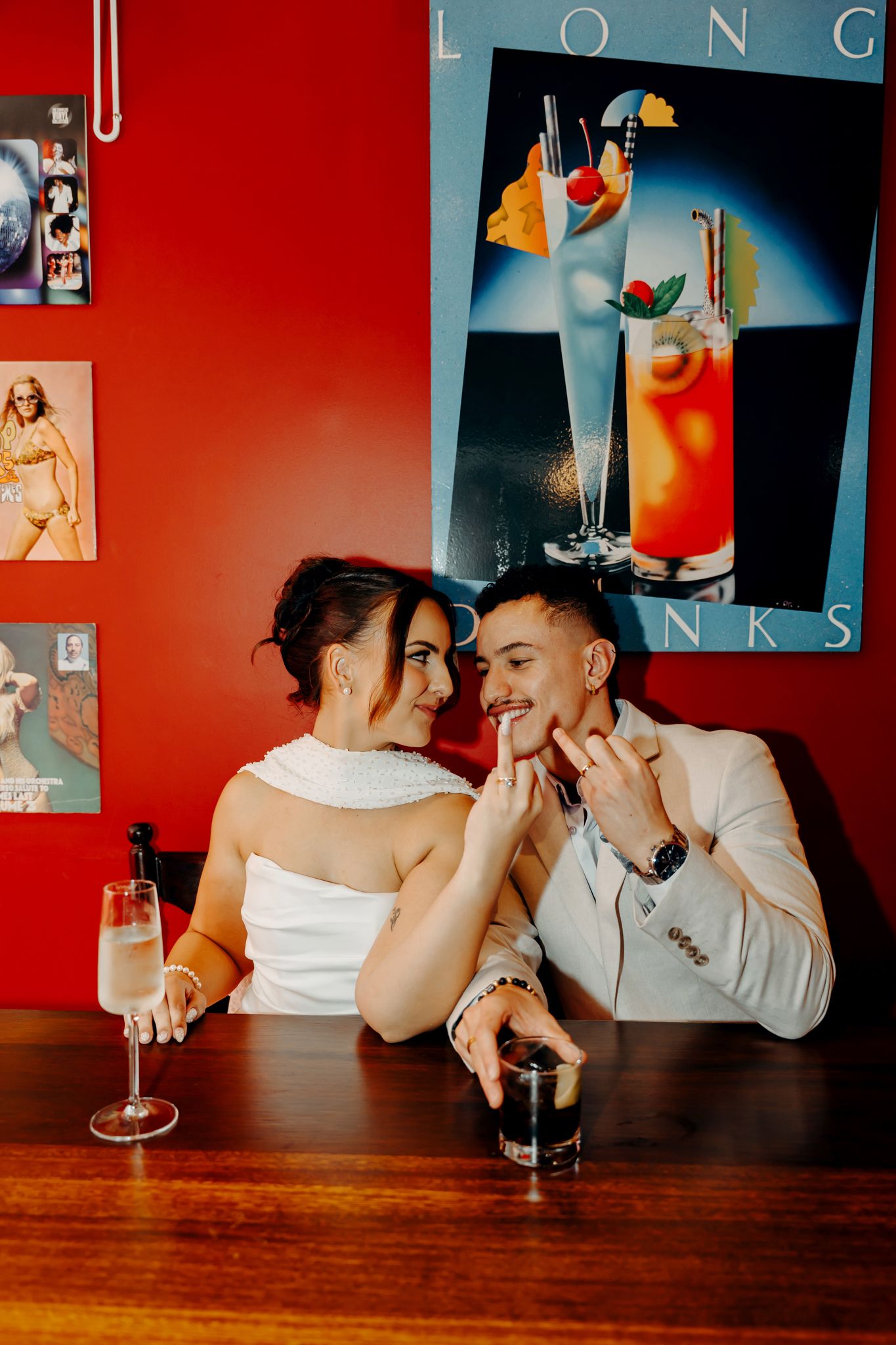 Couple in wedding attire sharing a romantic moment at a bar with cocktails and a colorful drinks poster on a red wall.