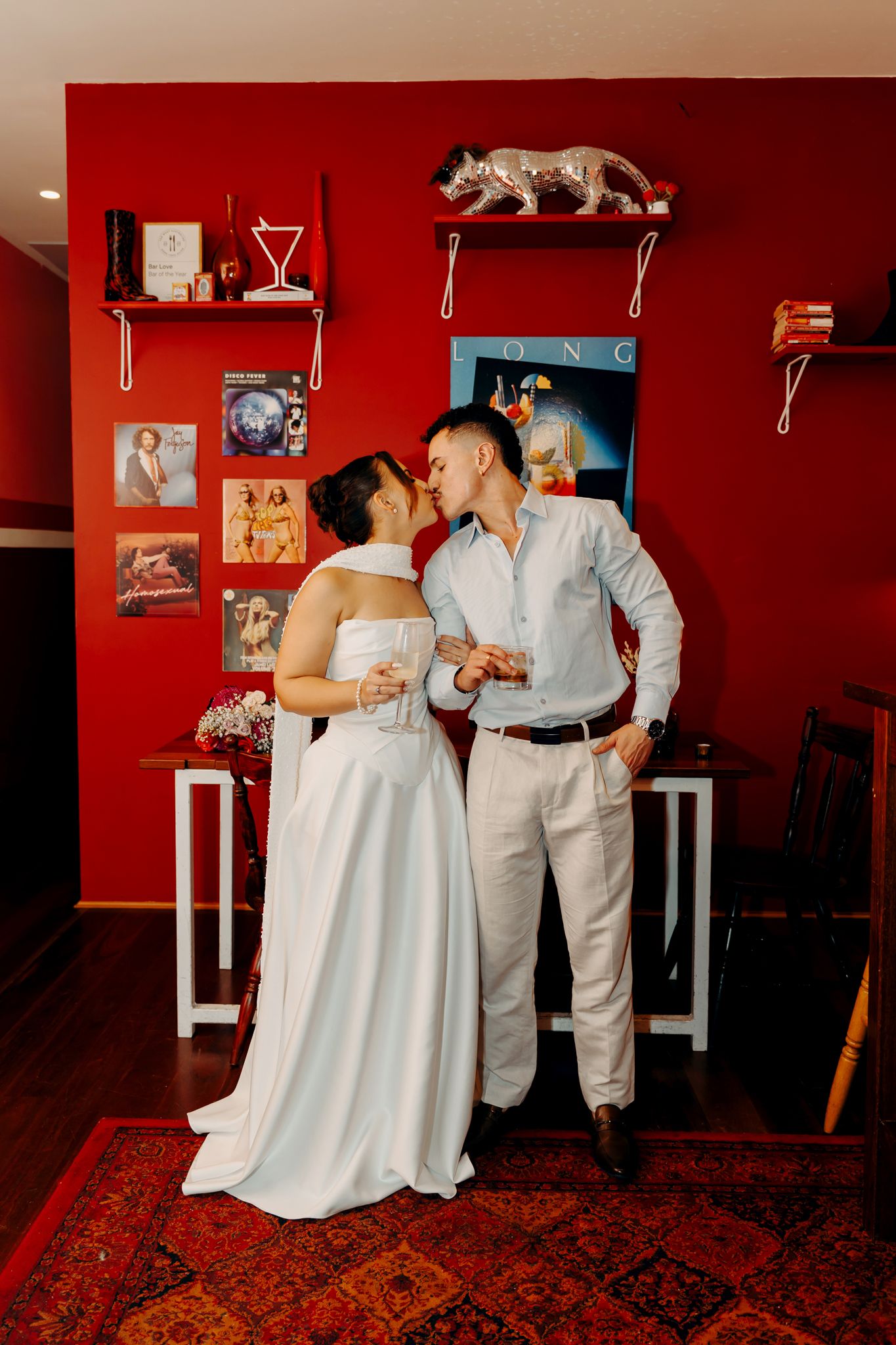 Wedding couple kissing while holding drinks against a bold red wall decorated with vinyl records and shelves.