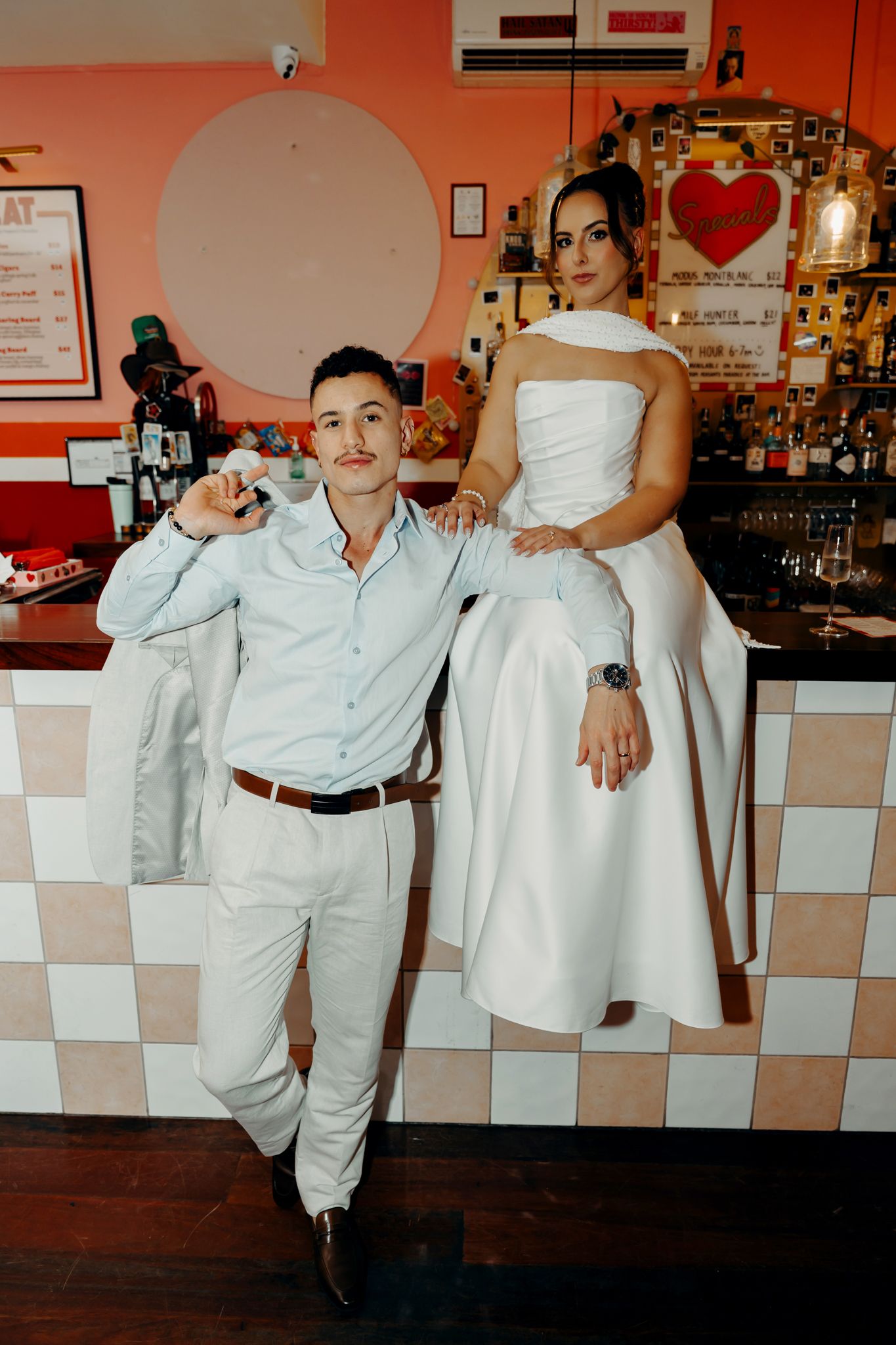 Bride in white dress sits on bar counter with groom in light blue suit at a retro-style restaurant.