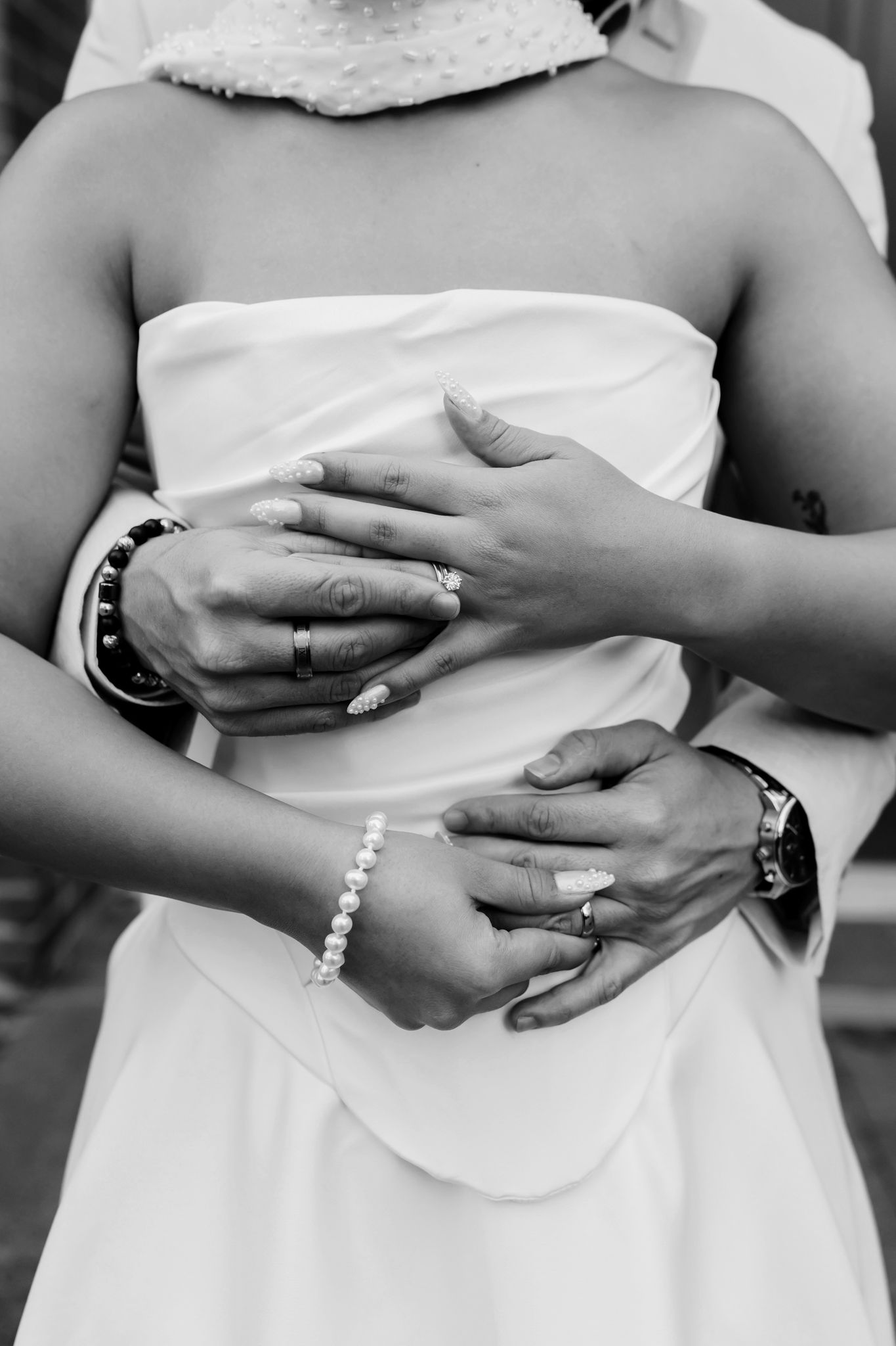 Black and white photo of bride and groom hands embracing, showing wedding rings and pearl bracelet.