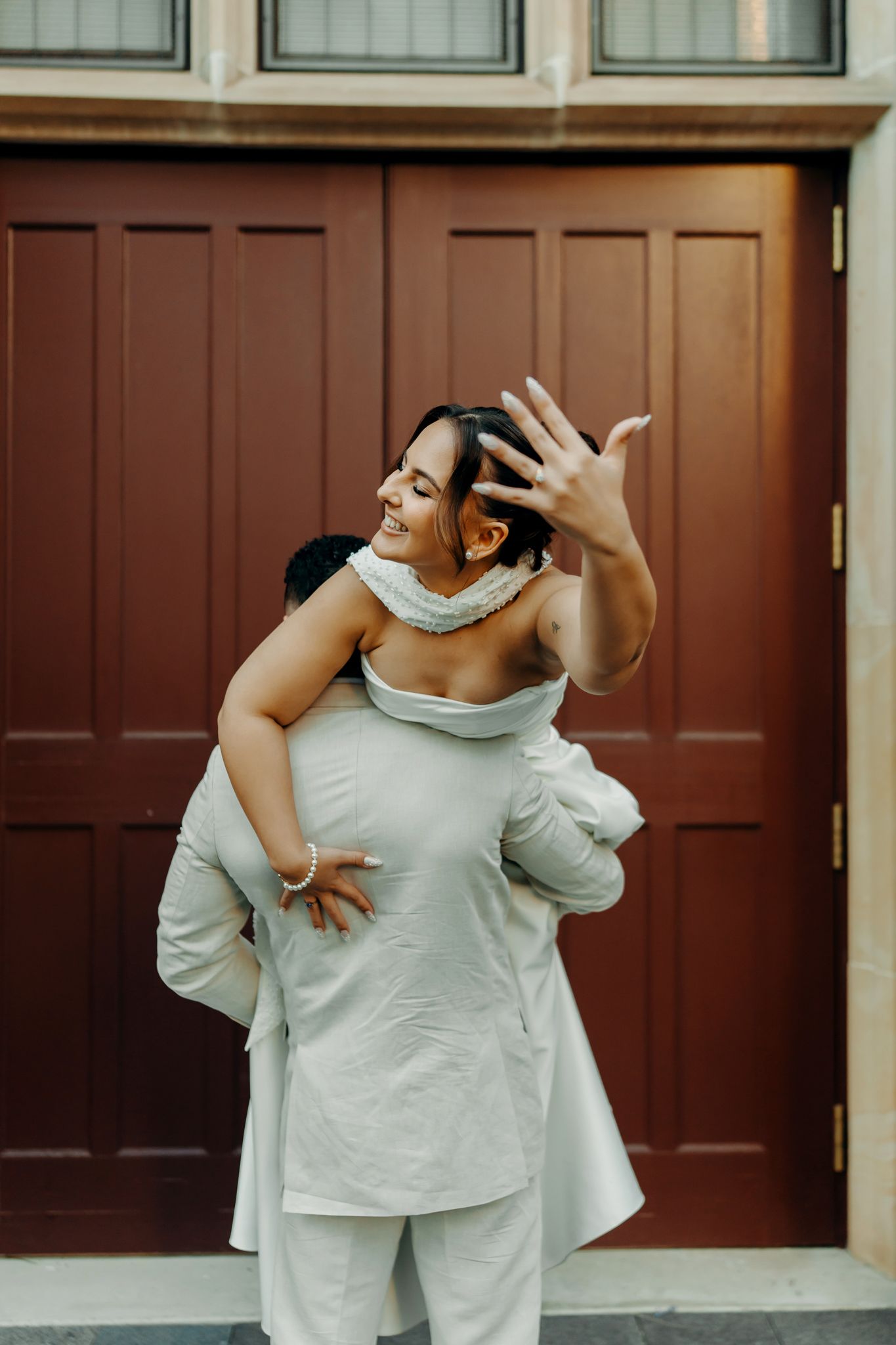 Groom carrying bride on his back outside a red door, bride smiling and showing off her wedding ring.