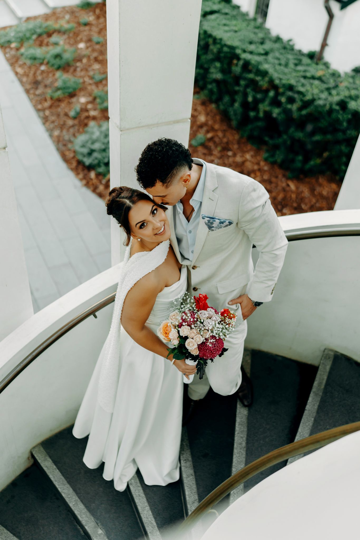 Bride and groom pose on spiral staircase, she holds colorful bouquet as he kisses her head in white wedding attire.