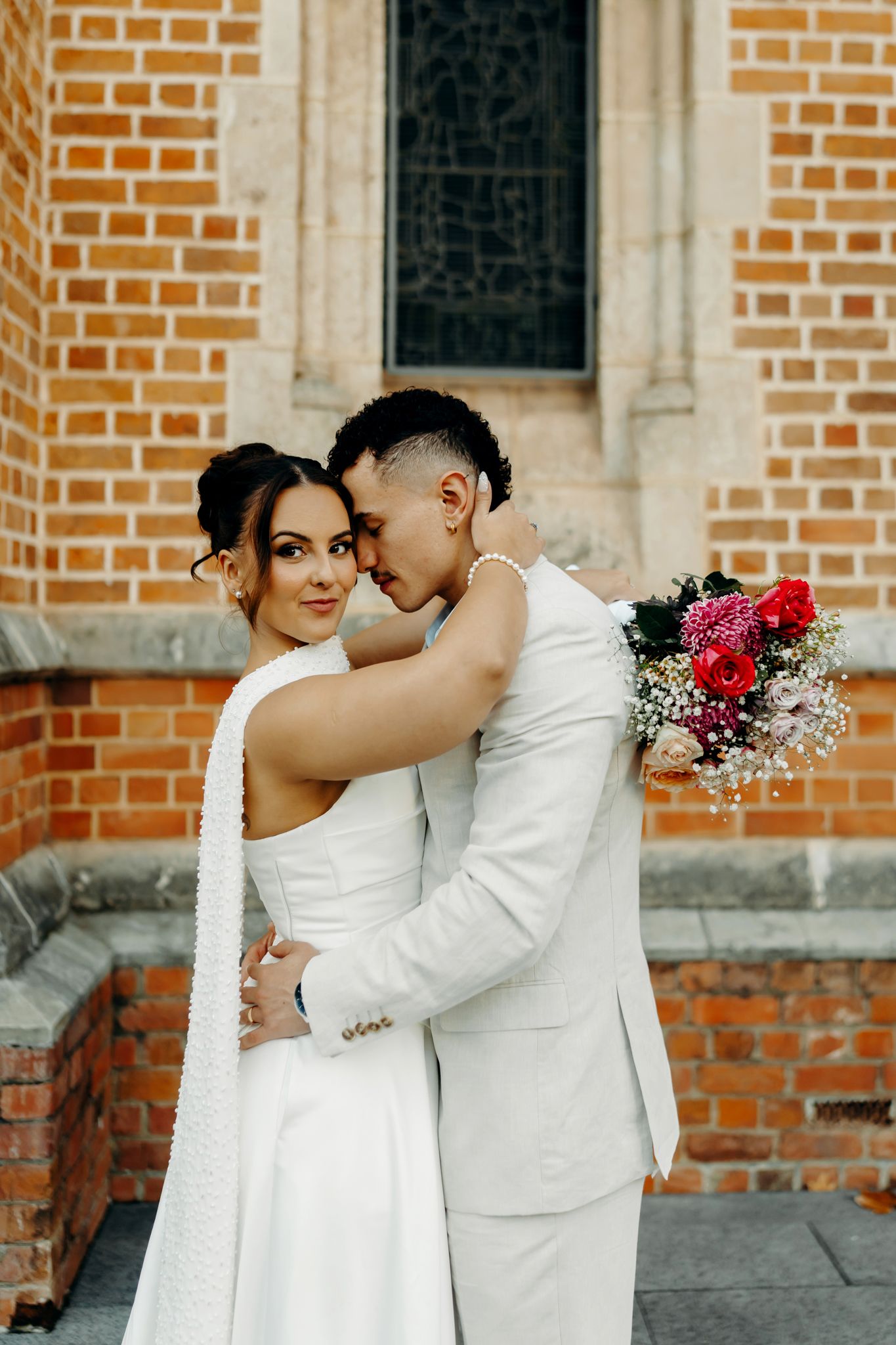 Bride and groom embracing in front of a brick church, bride holding a colorful bouquet, both dressed in white.
