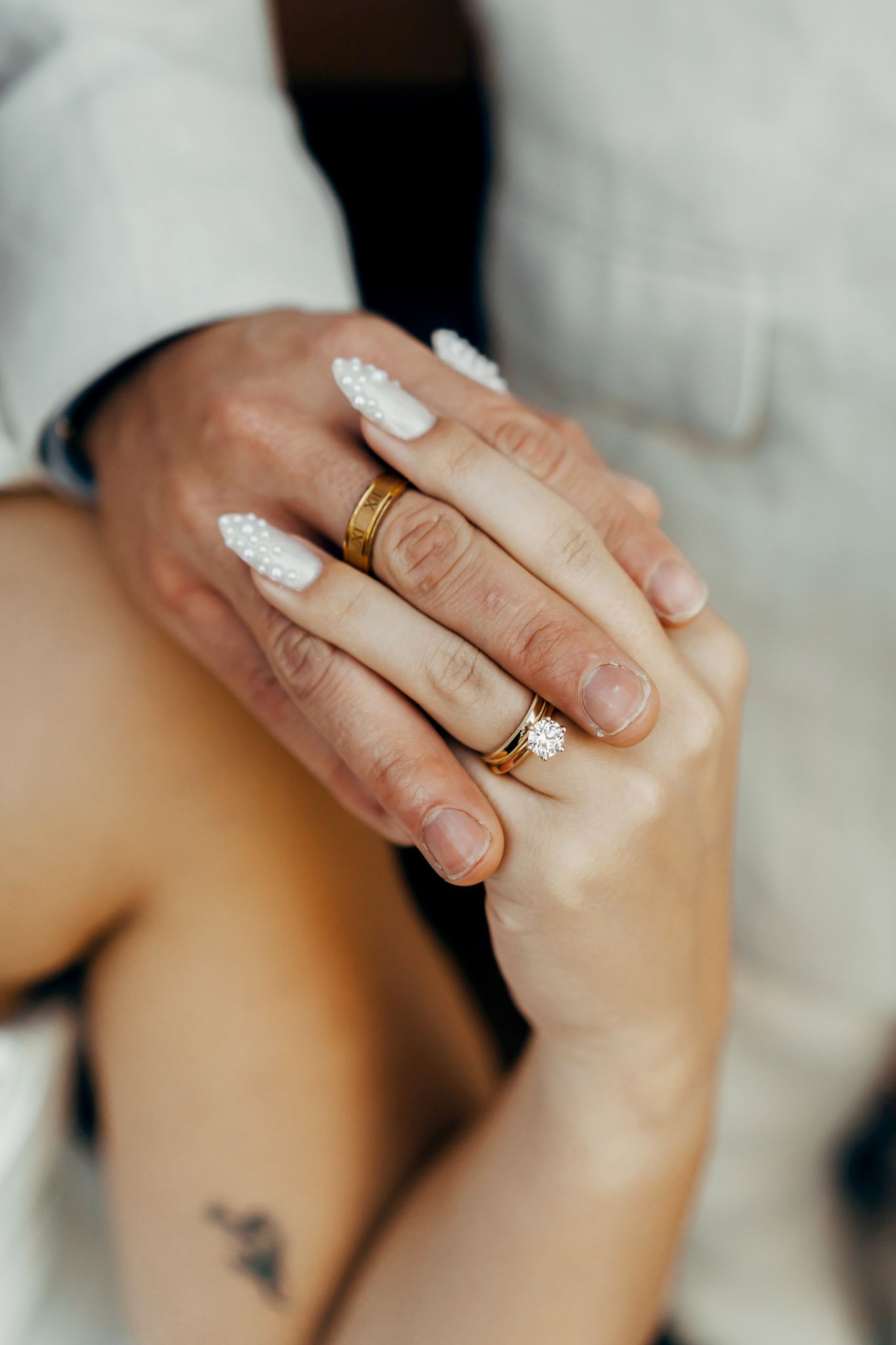 Close-up of couple holding hands showing matching gold wedding bands, with white nail art and a small tattoo visible.