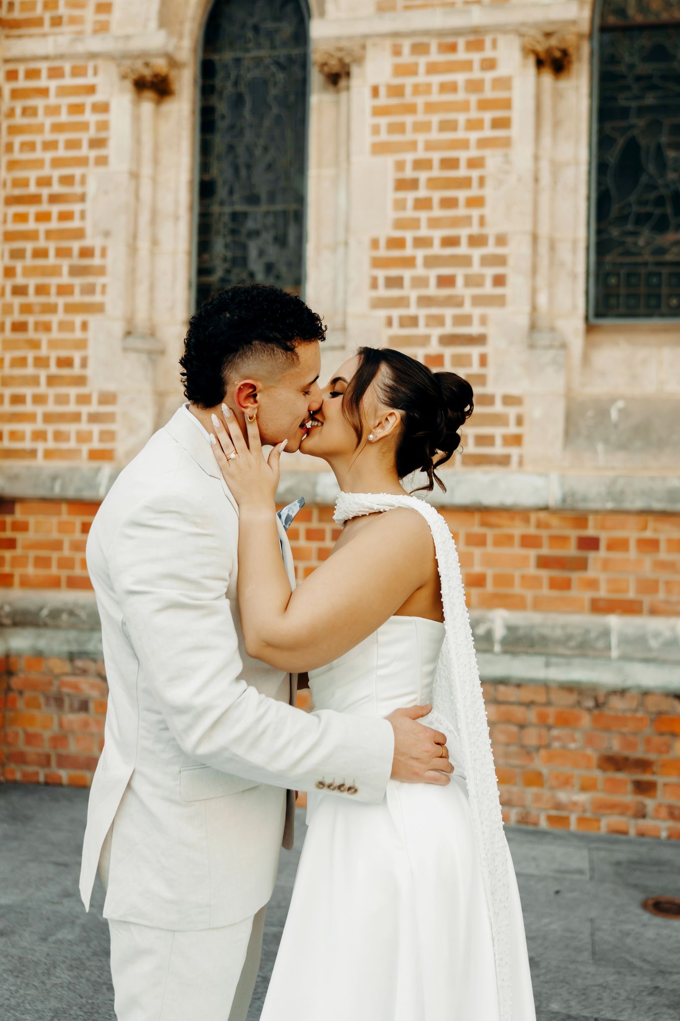 Newlyweds share a romantic kiss outdoors in front of a rustic brick wall building.