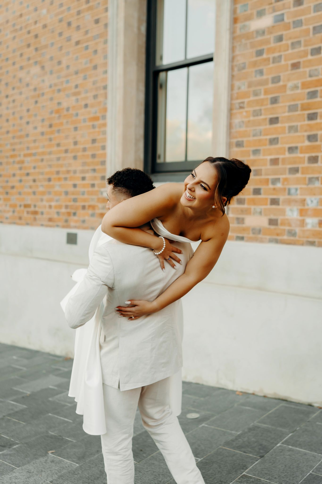 Joyful bride in white strapless gown being lifted by groom in white suit outside brick building.