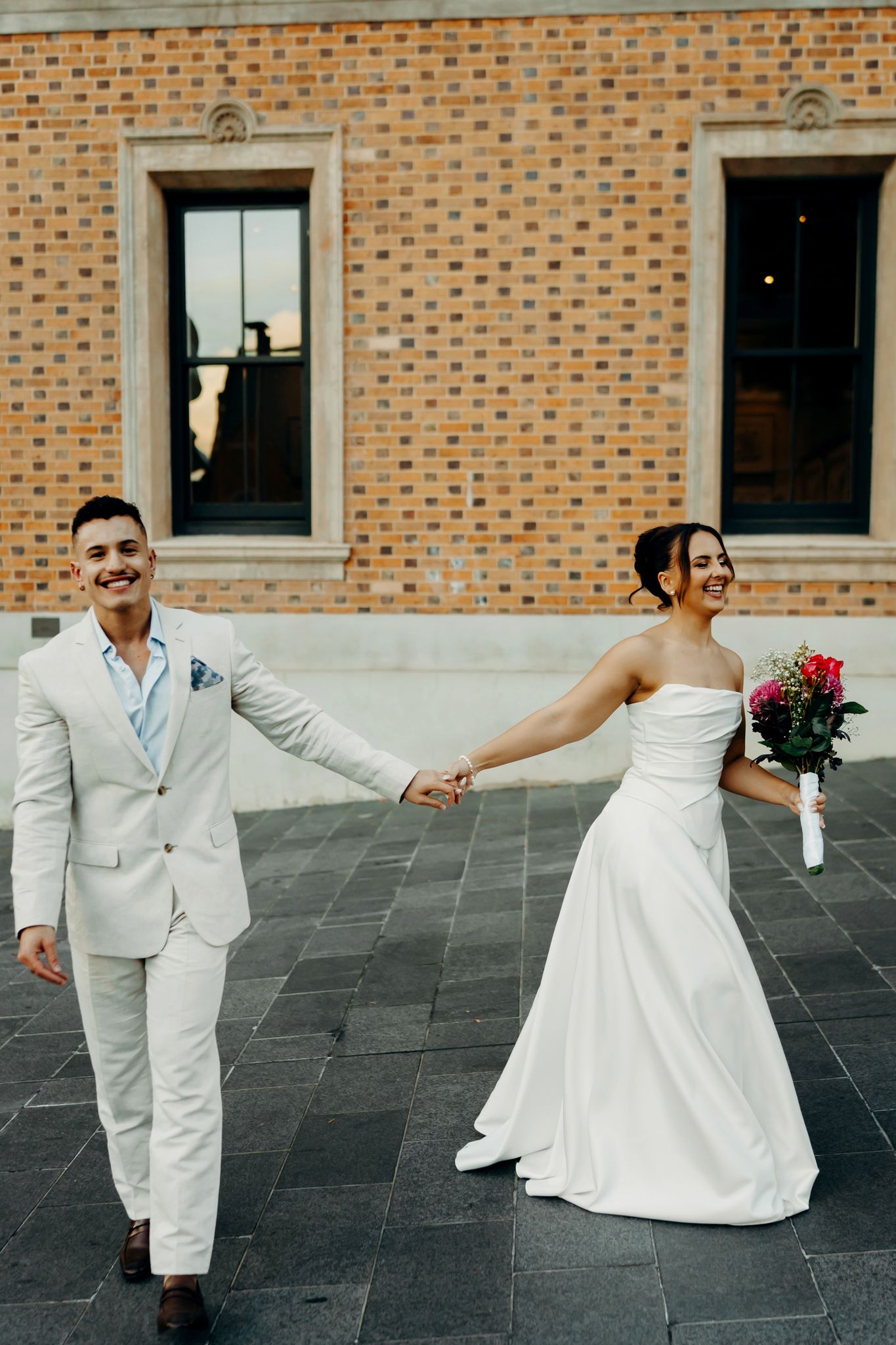 Smiling bride and groom in white wedding attire holding hands walking outside brick building.