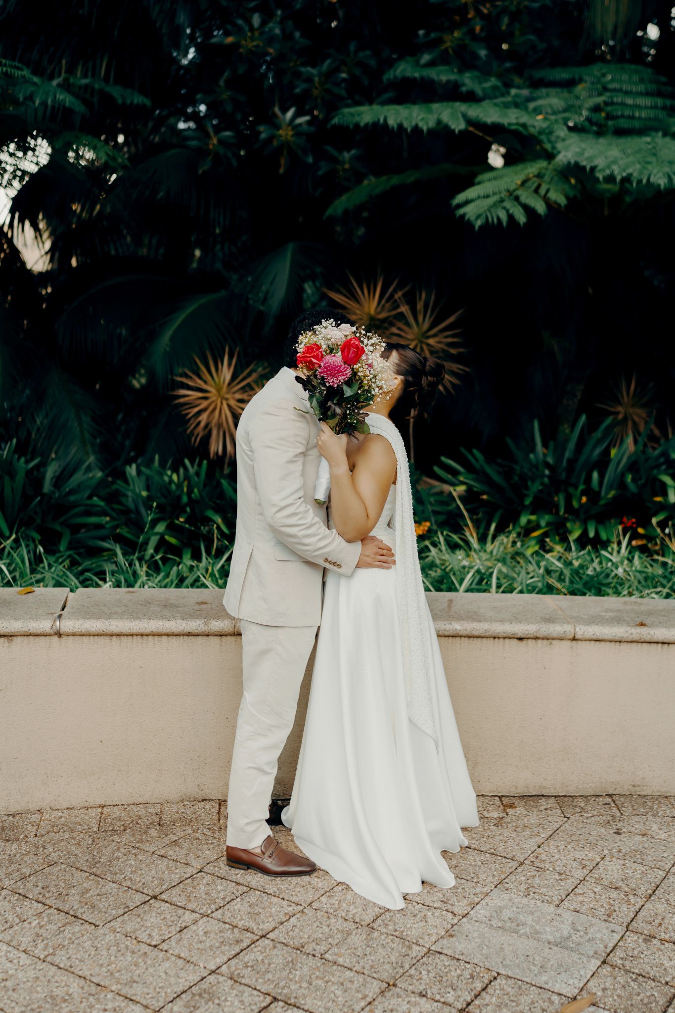 Bride and groom sharing a kiss while holding a colorful bouquet in front of lush tropical garden greenery.
