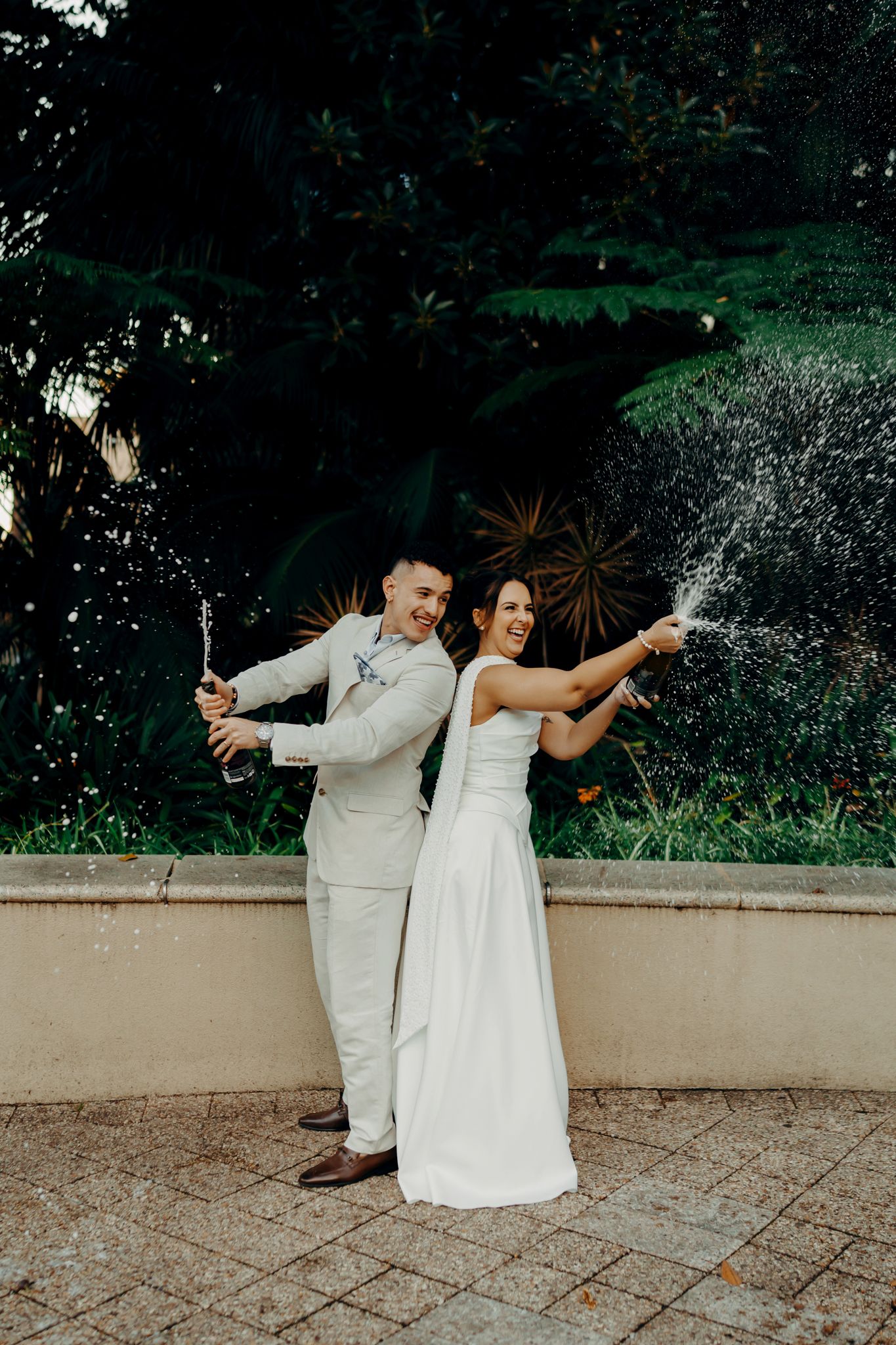 Joyful newlyweds popping champagne bottles back-to-back outdoors with tropical plants in the background.