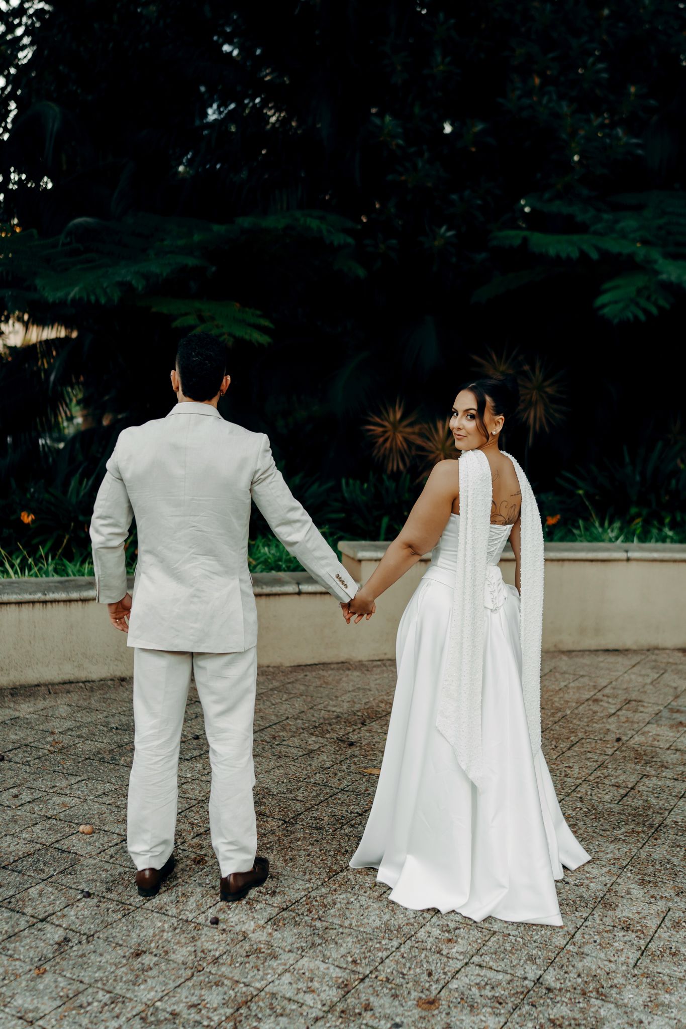 Bride and groom holding hands from behind, standing before lush green garden in wedding attire.