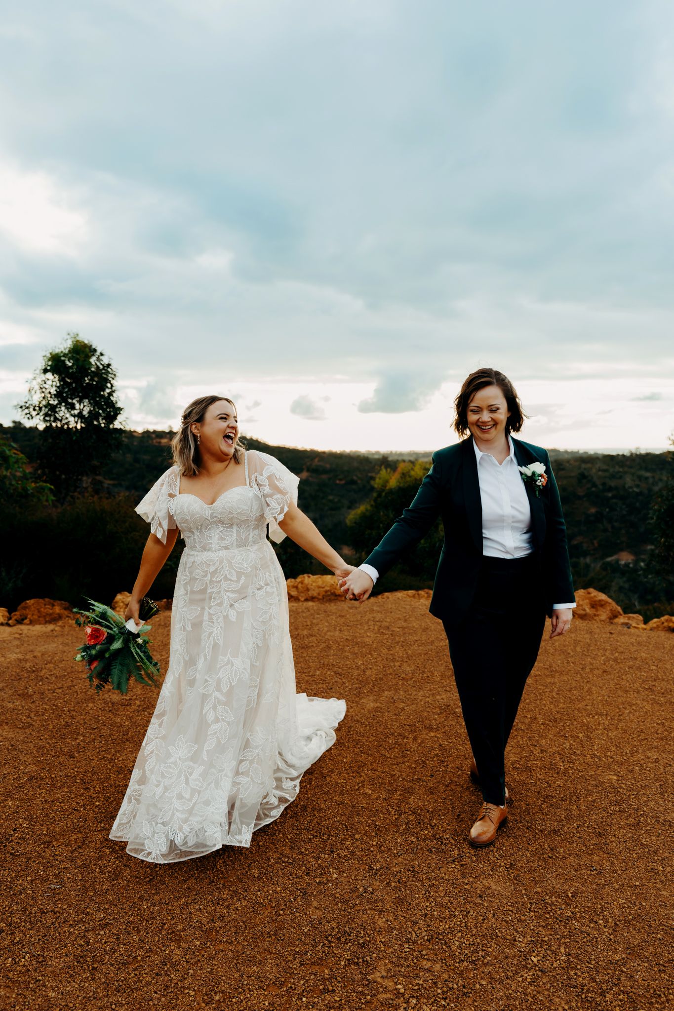 A couple in wedding attire walks together on a rustic dirt path with a cloudy sky and trees in the background.