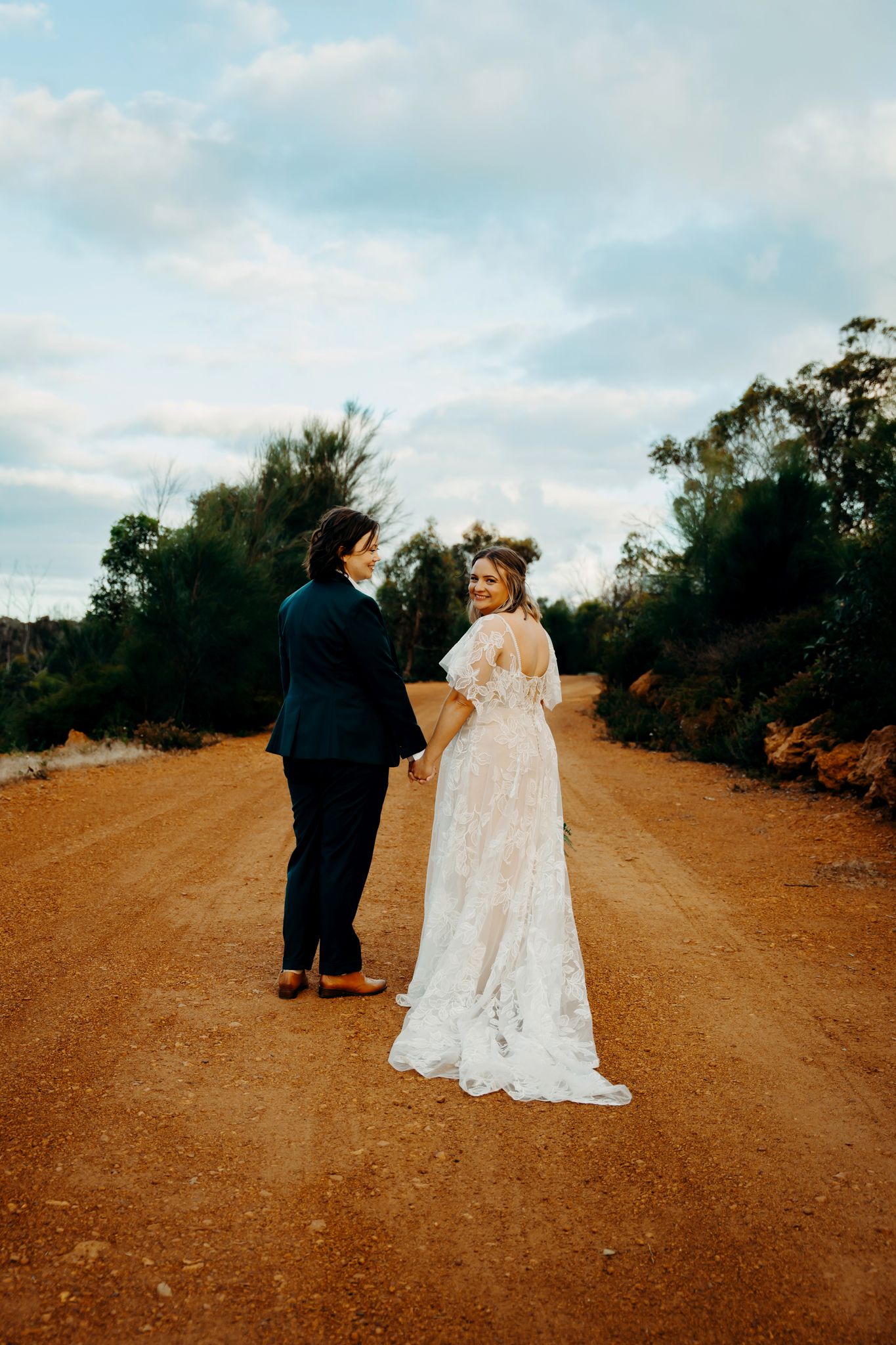 A couple in wedding attire walks together on a rustic dirt road surrounded by desert brush and trees.