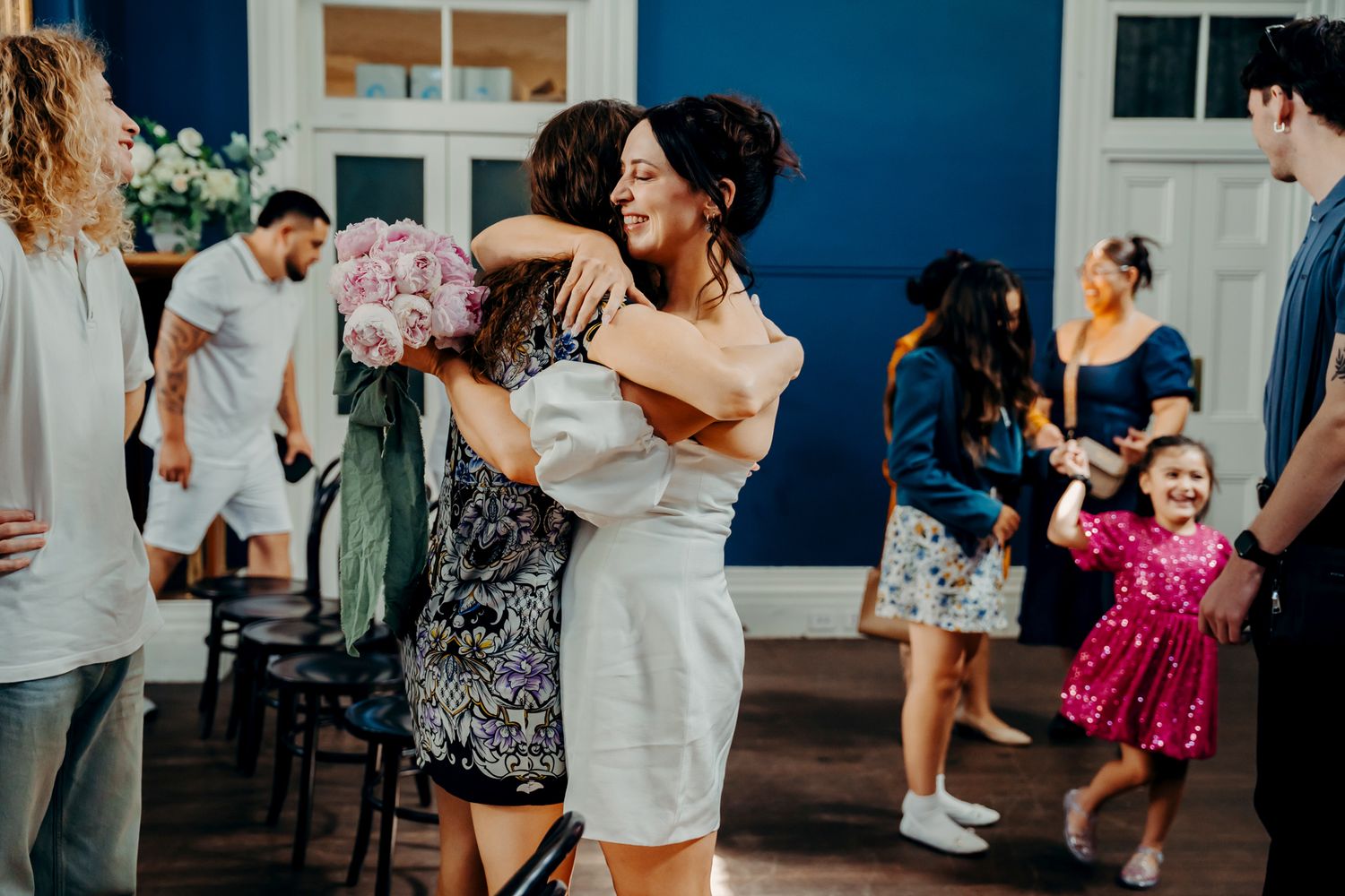 People dancing and embracing at a casual gathering with a bright blue wall in the background while others mingle.