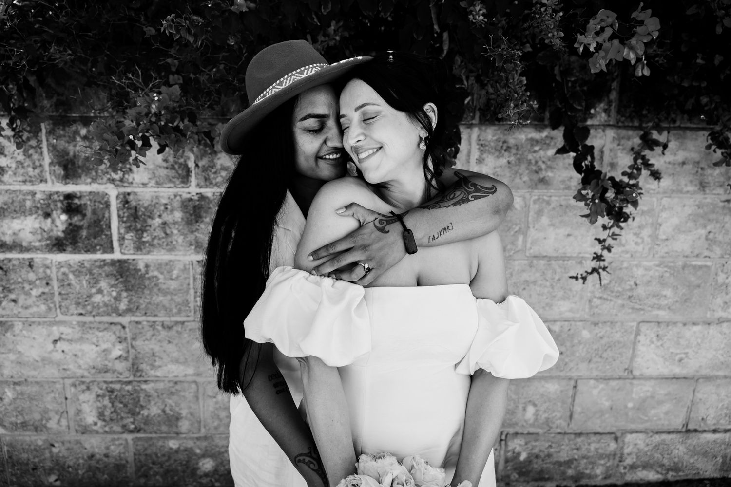 Black and white photo showing two people embracing and smiling while standing against a stone wall.