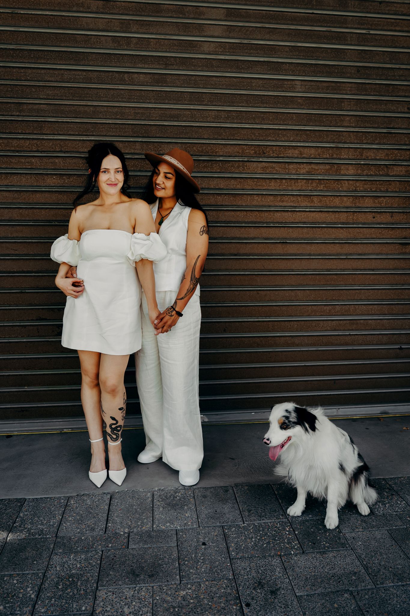 Two people in white outfits sit together on stone steps with their fluffy white dog against an industrial backdrop.