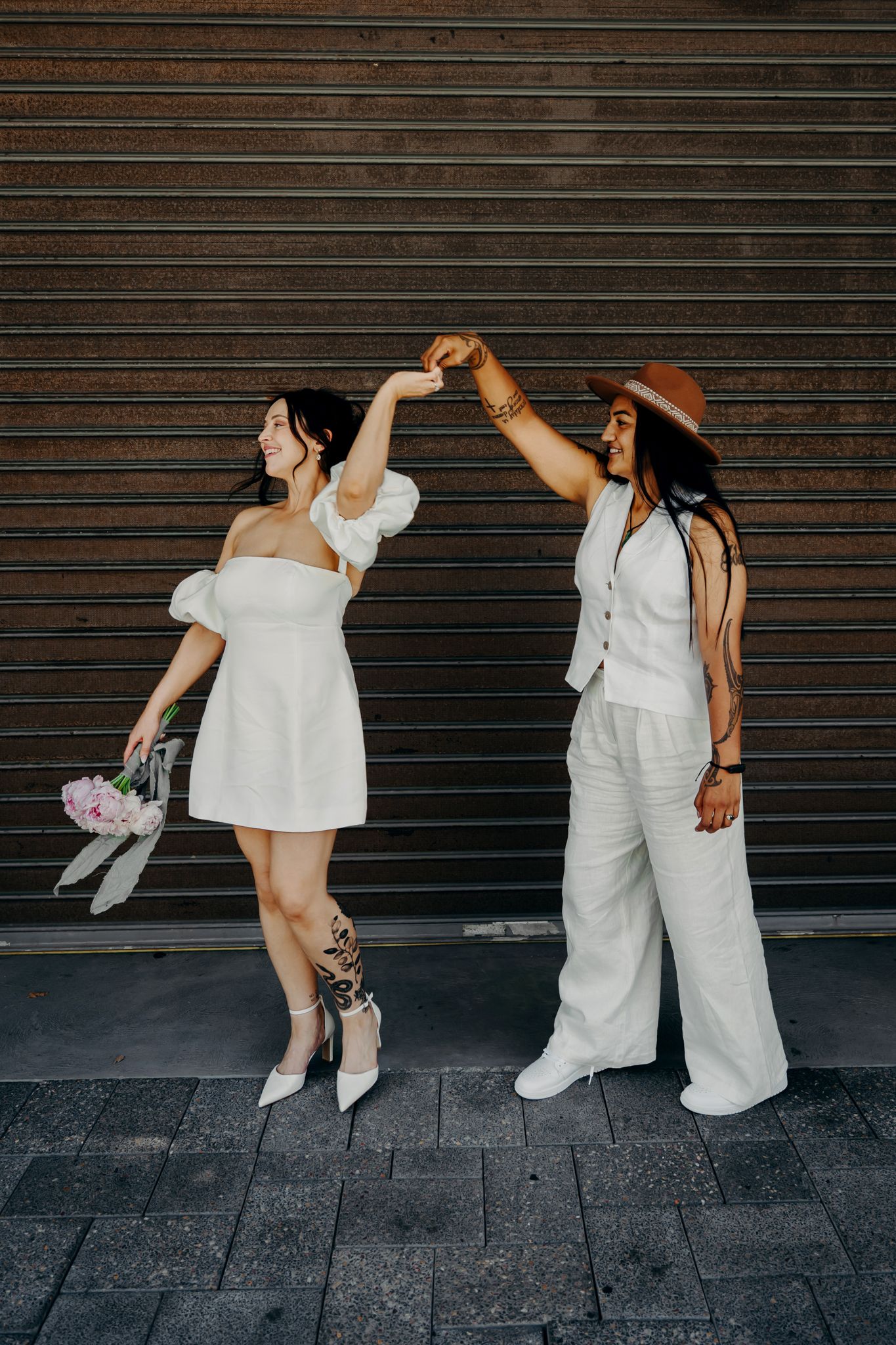Two people dancing together in coordinated white outfits against an industrial metal wall background.