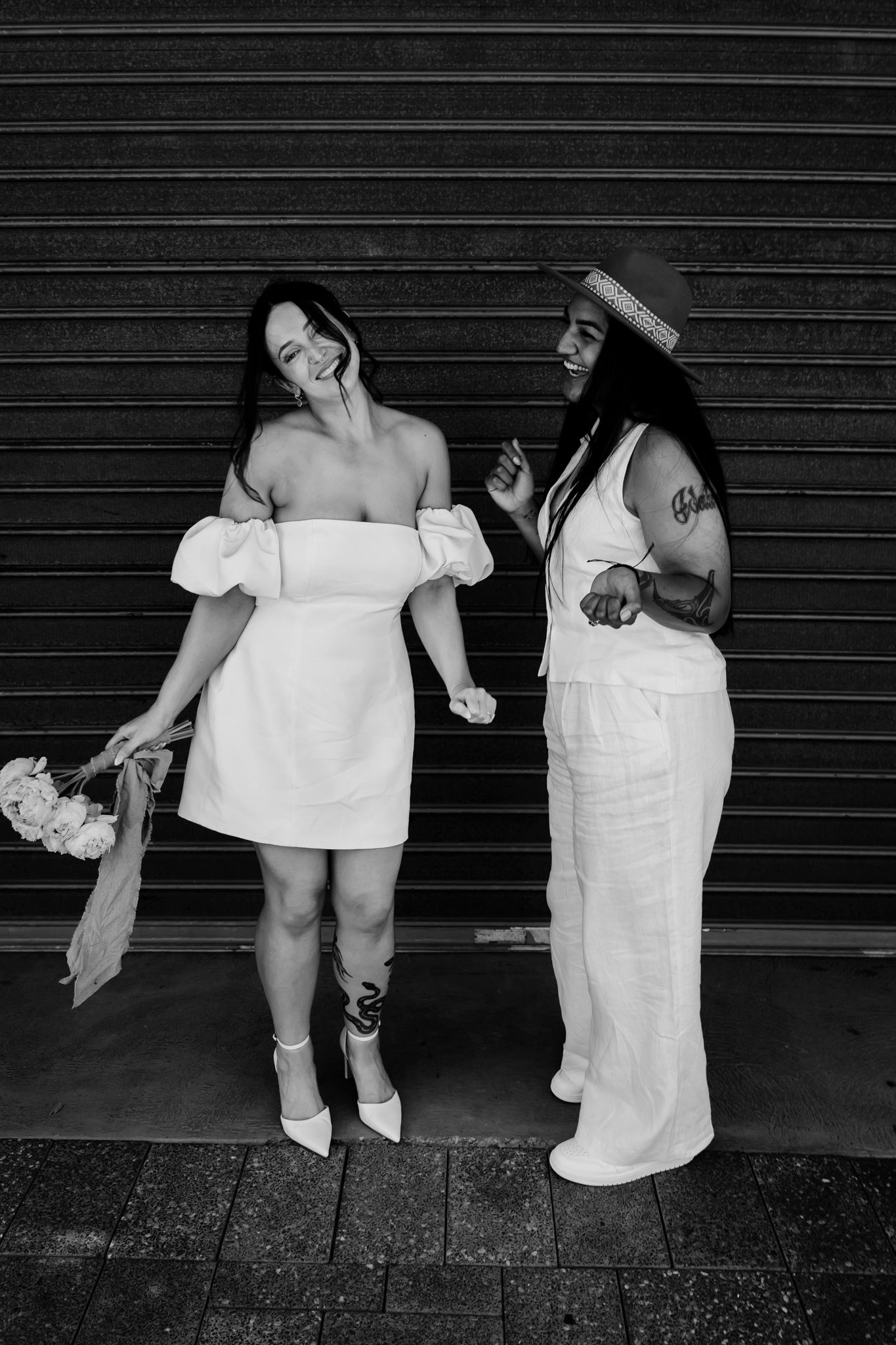 Two people in white dresses share a candid moment against a black striped wall, both holding bouquets and wearing summer attire.
