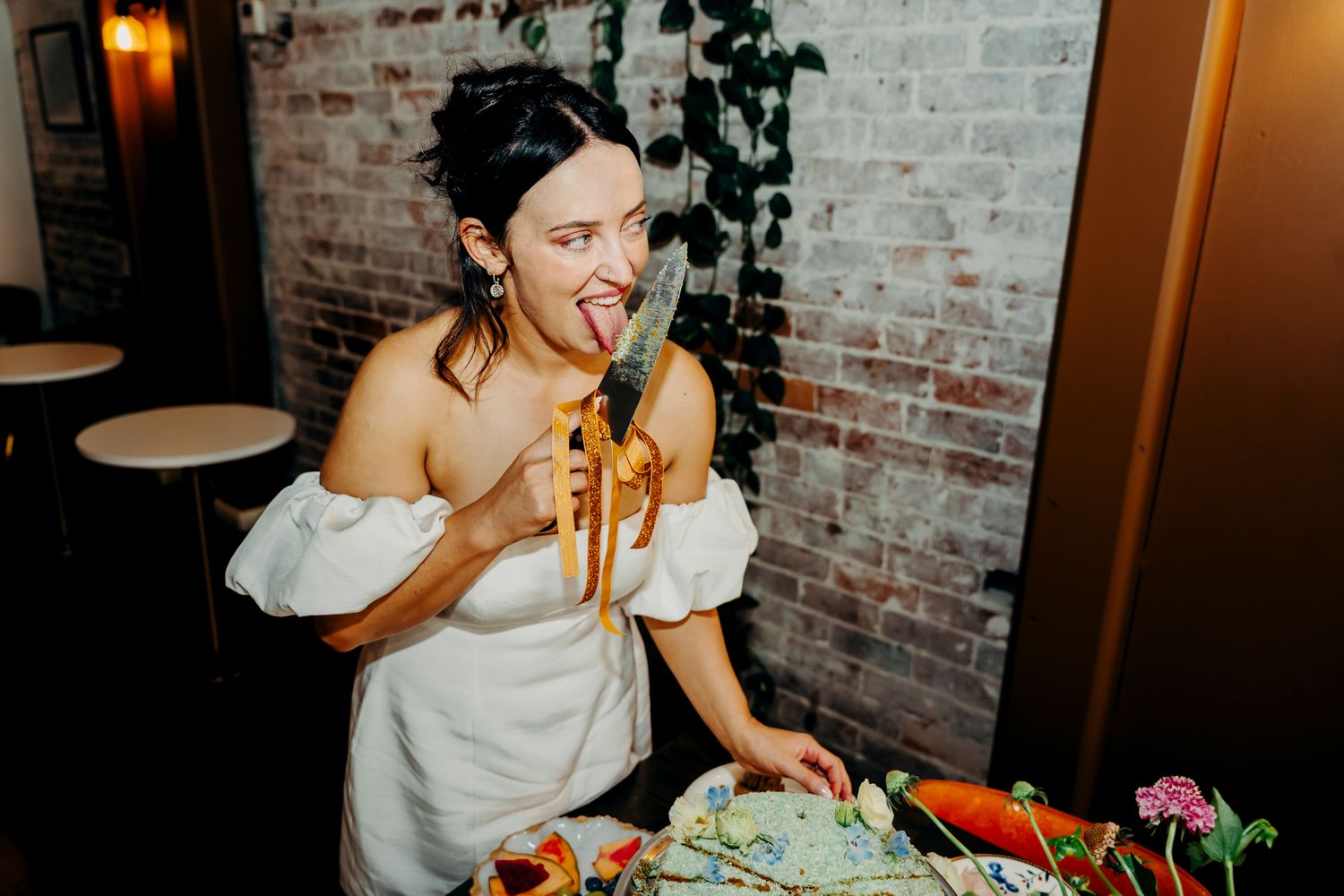 Someone in a white off-shoulder dress enjoys eating from a table of appetizers at a brick-walled venue.