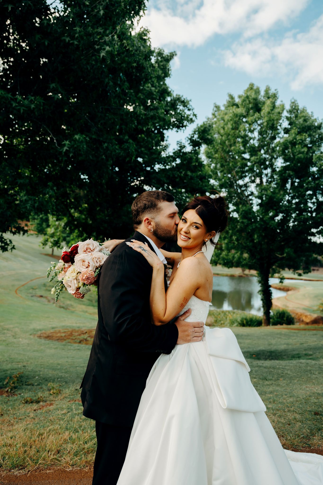 A groom in a black tuxedo kisses his bride in a white ballgown outdoors, as she smiles holding a pink floral bouquet.