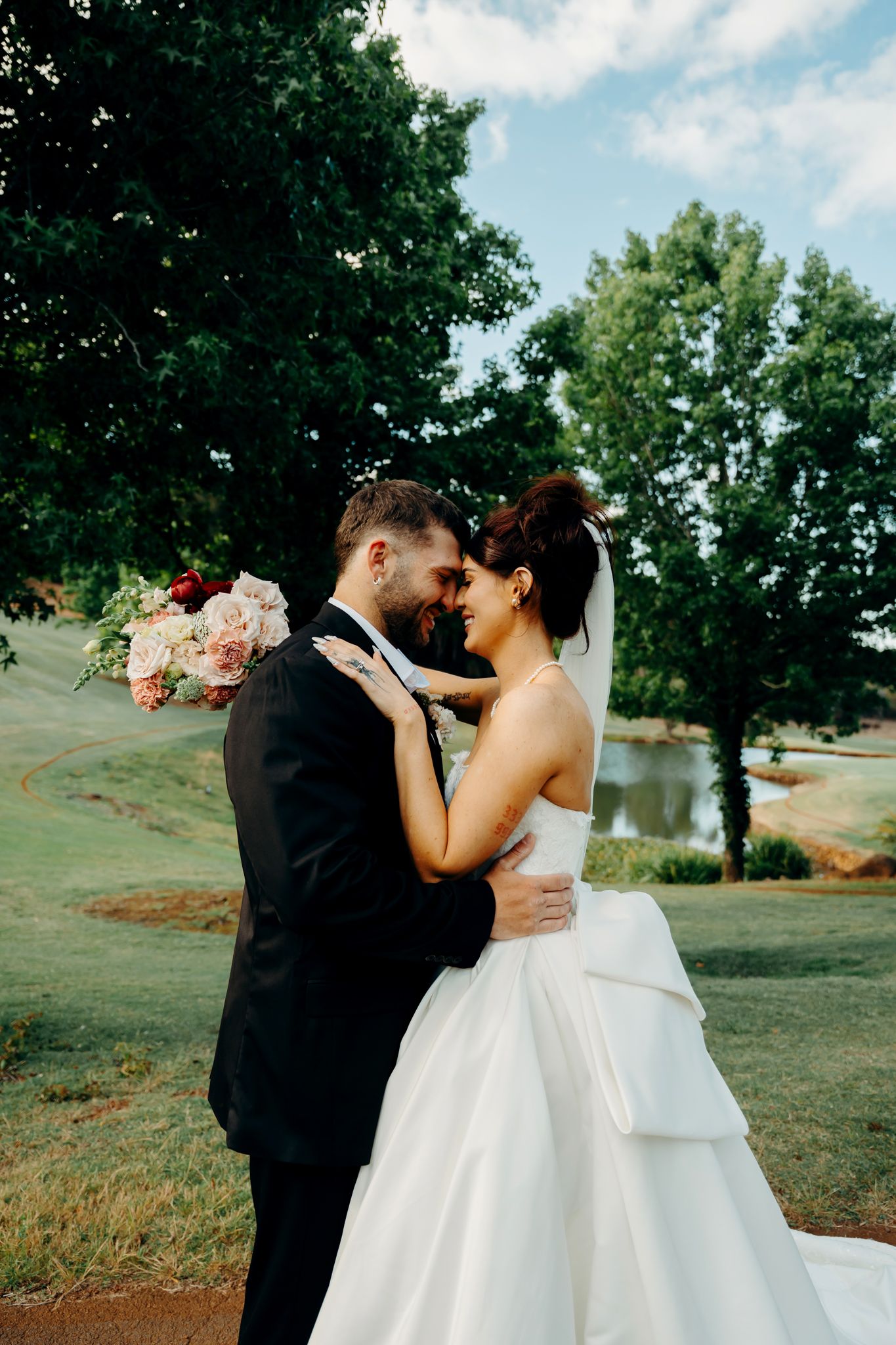 Bride and groom share a romantic forehead touch outdoors, with lush green trees and a pond in the background.