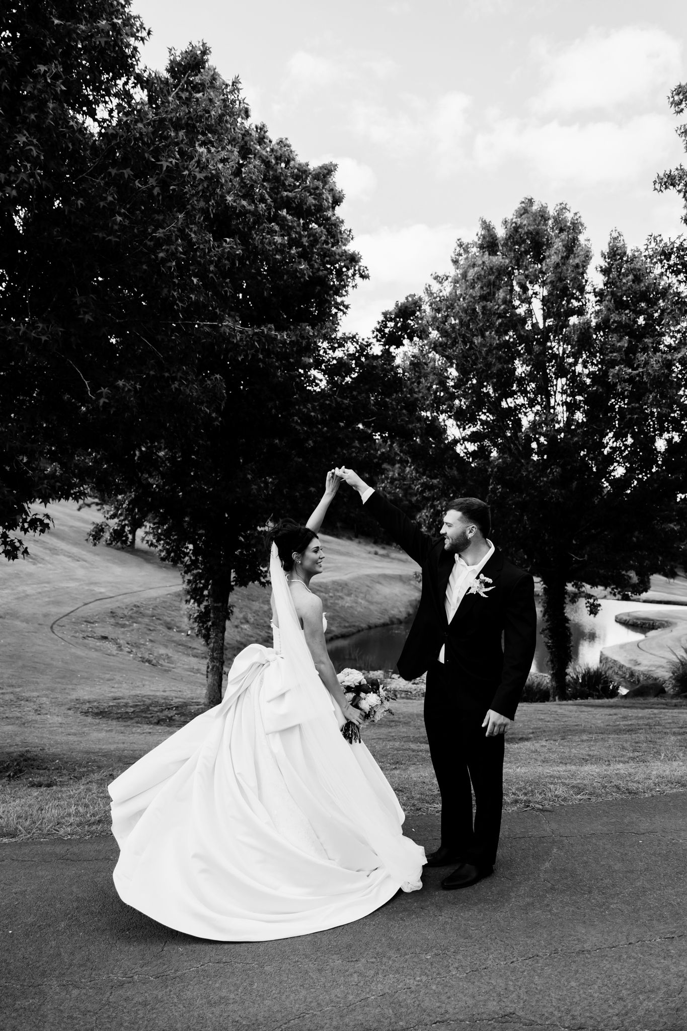 Black and white photo of bride and groom dancing outdoors, bride raising bouquet, flowing white gown, trees in background.