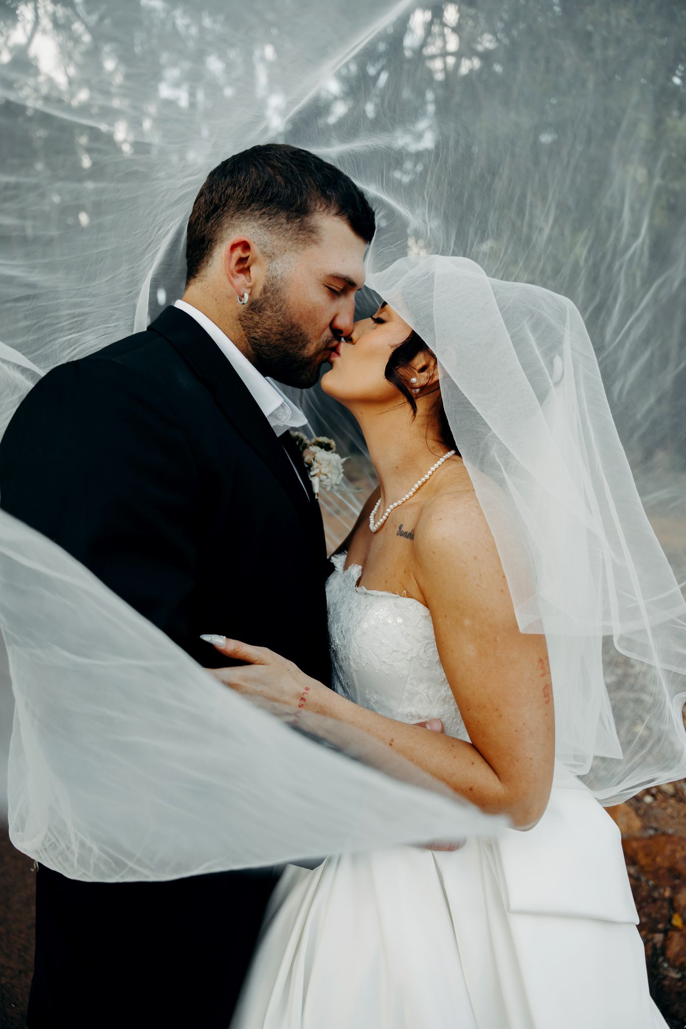 Bride and groom share a kiss under a flowing veil, creating an ethereal and romantic wedding portrait.
