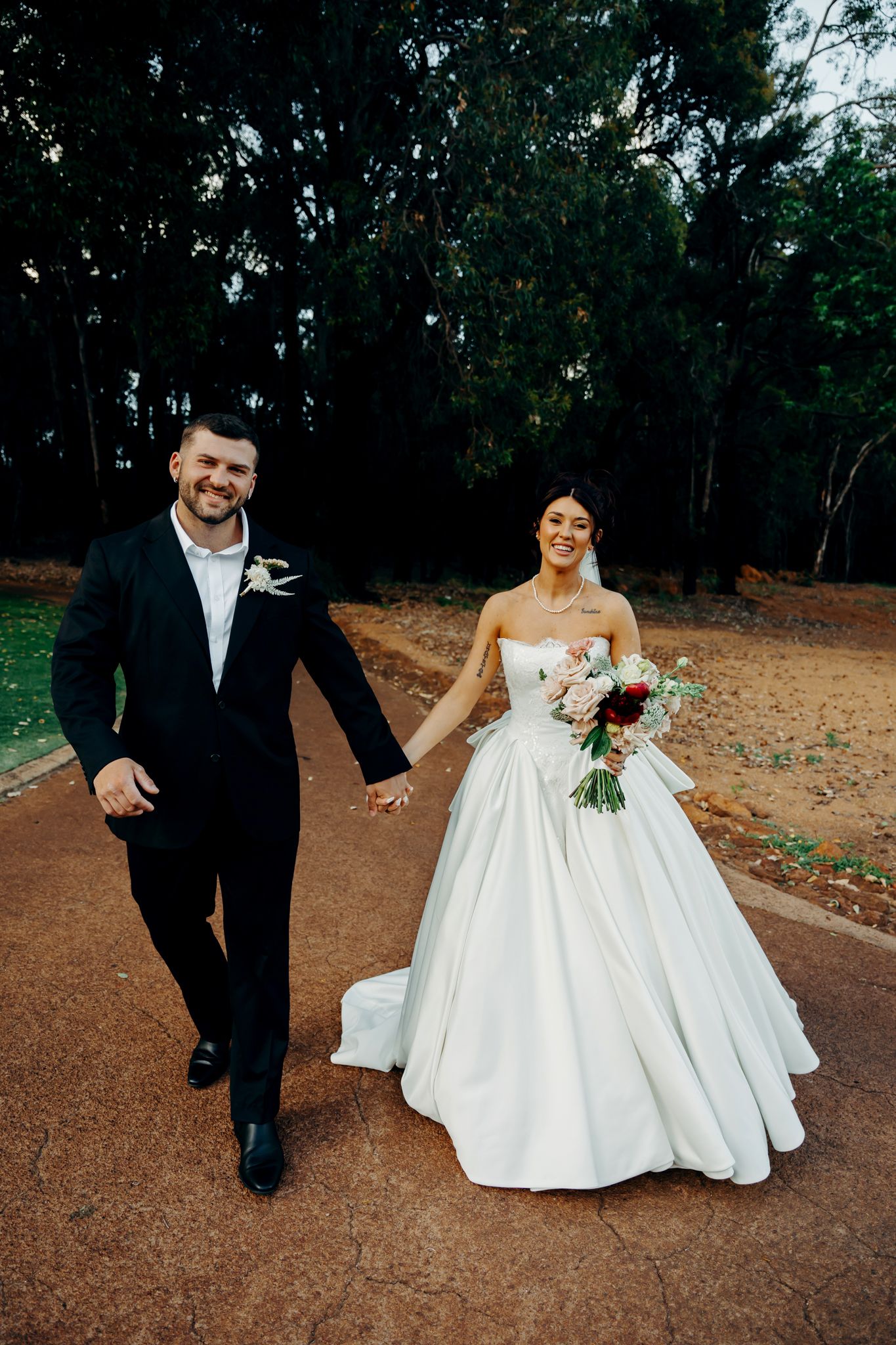 Bride in white ballgown and groom in black suit walk hand in hand on a path surrounded by trees.