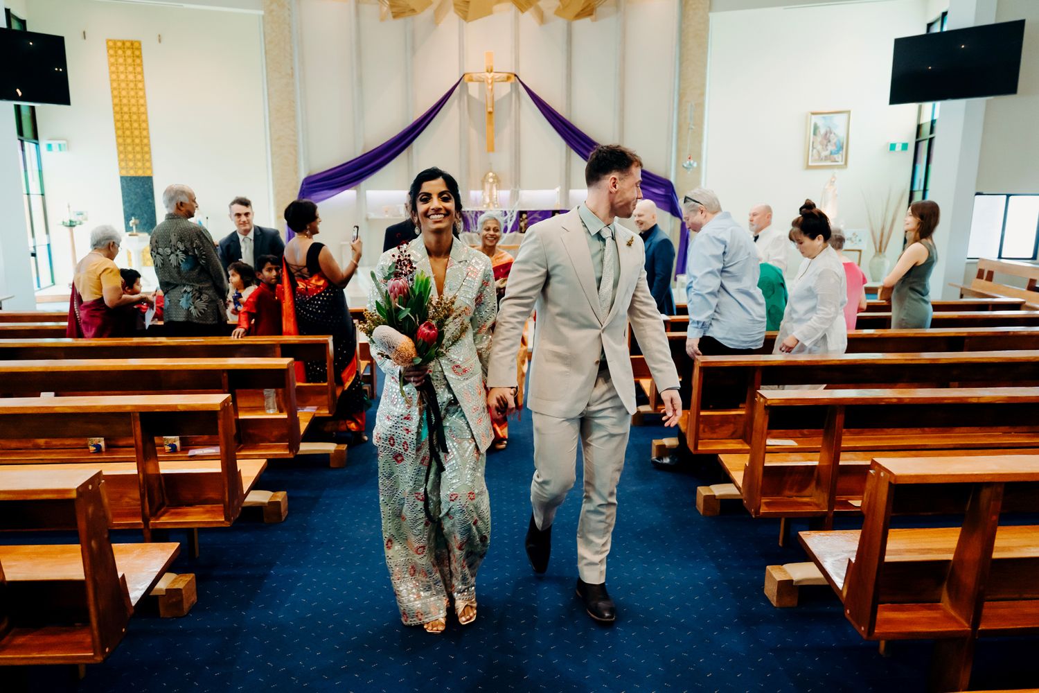 A couple walks down the aisle in a small chapel decorated with purple fabric, surrounded by wooden pews and guests.