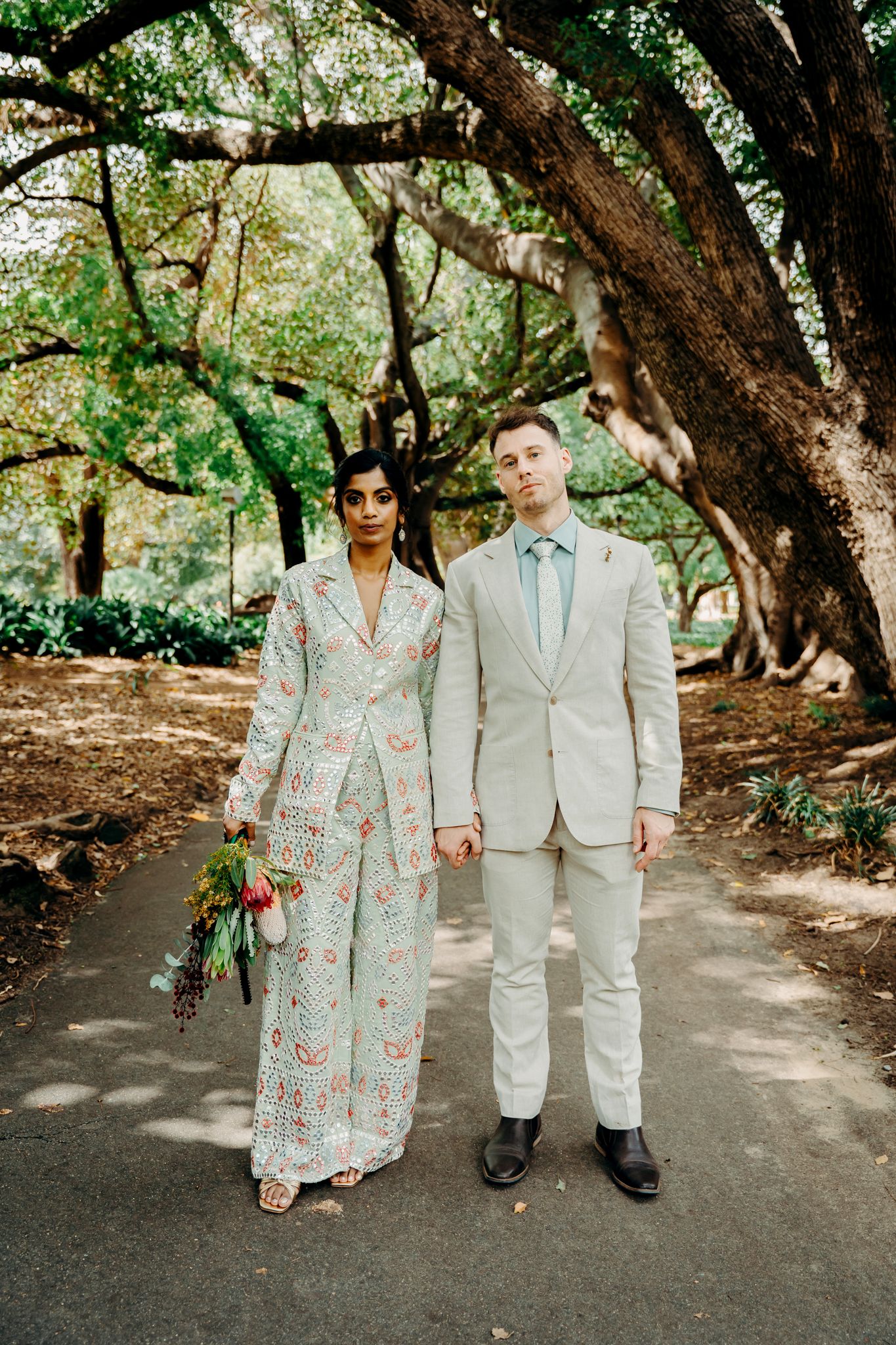 A couple in coordinated wedding attire poses together beneath lush green trees on a park path.