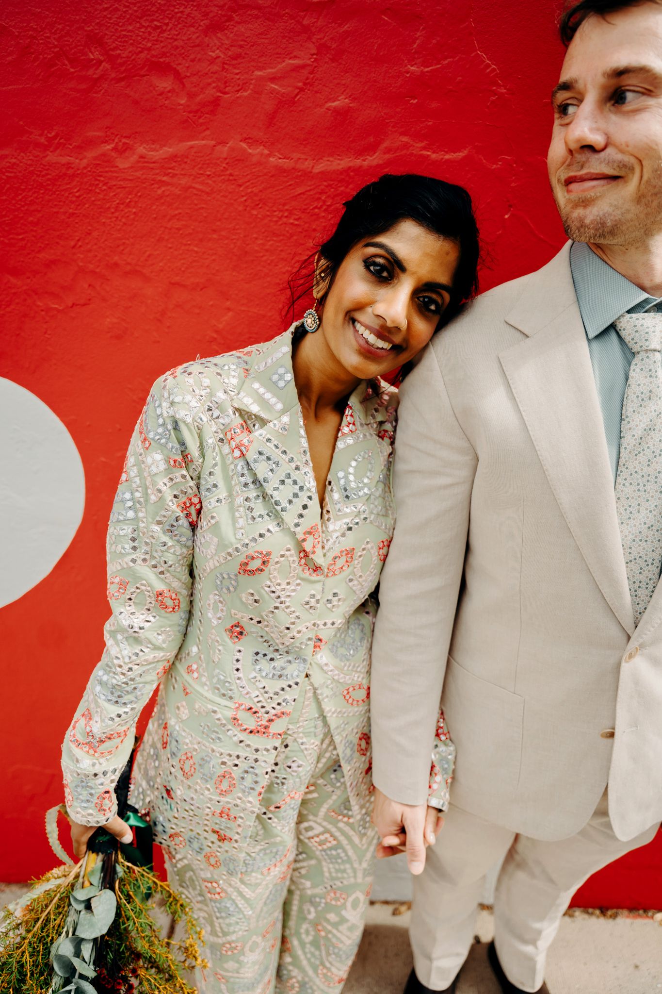 A wedding couple in a floral print dress and beige suit pose playfully against a vibrant red wall background.