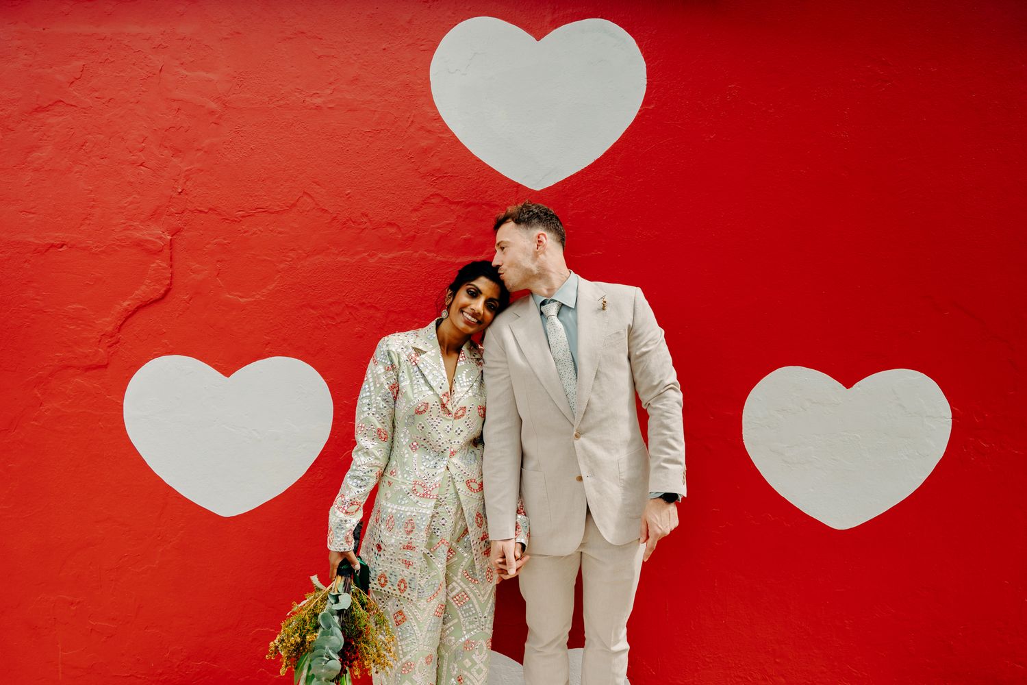 Romantic wedding couple stands against vibrant red wall with three white painted hearts as backdrop.