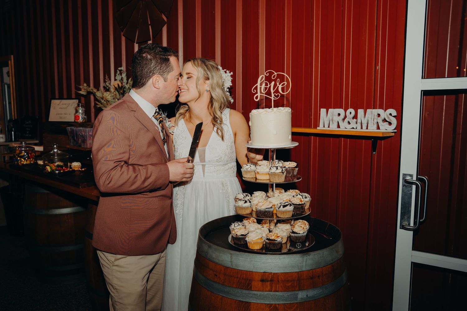 Couple sharing a kiss by wedding cupcake display on rustic wine barrel stand.