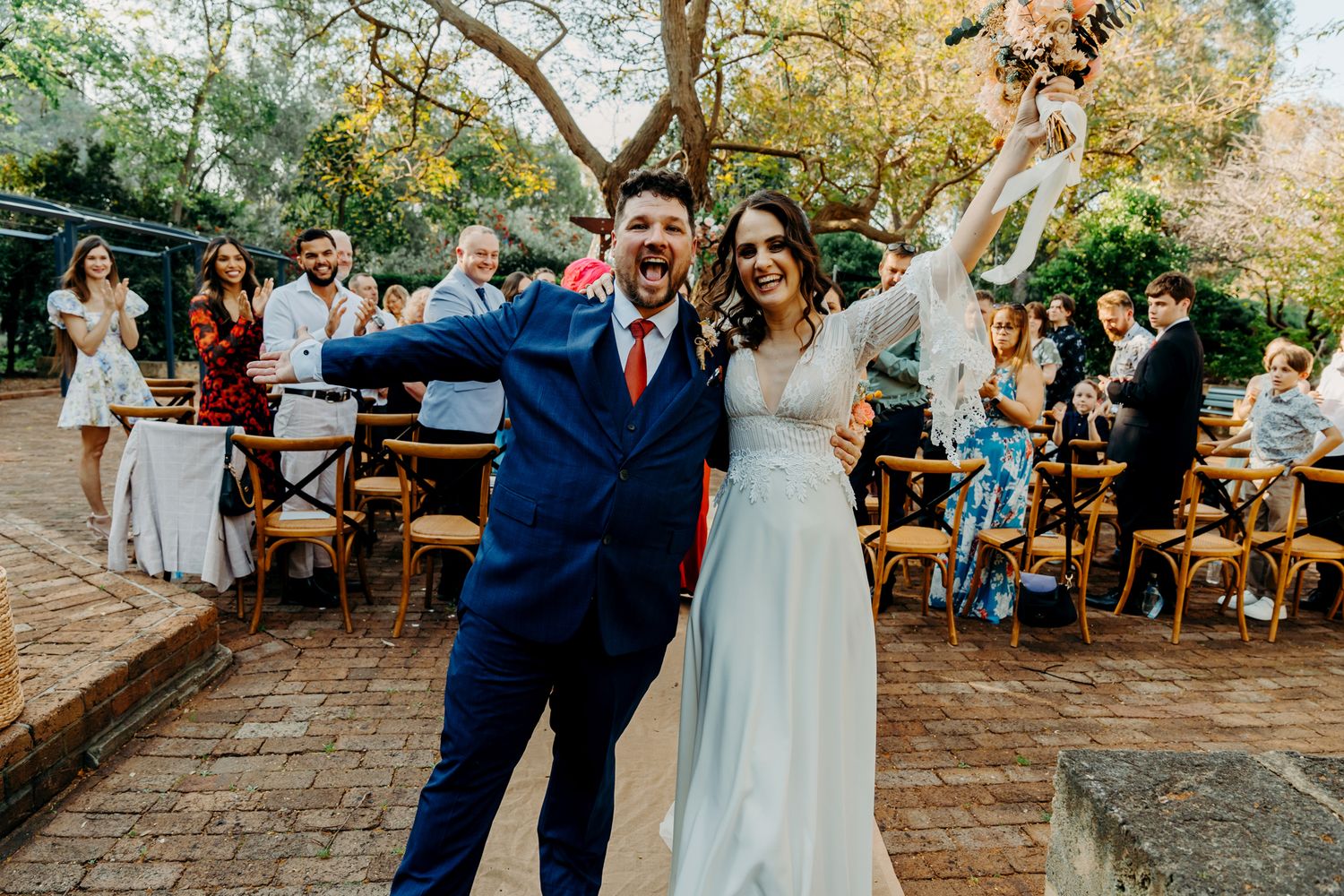Wedding couple celebrates joyfully at an outdoor reception with rustic wooden chairs and natural decor.