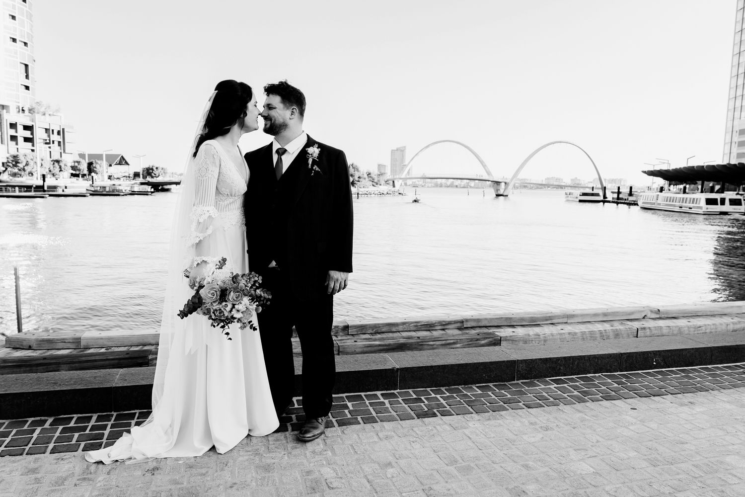 A romantic couple embraces on a dock by the water with a bridge visible in the distant background at sunset.