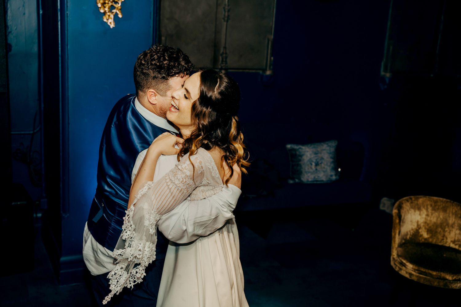 A couple shares an intimate embrace in a dimly lit room with royal blue walls.
