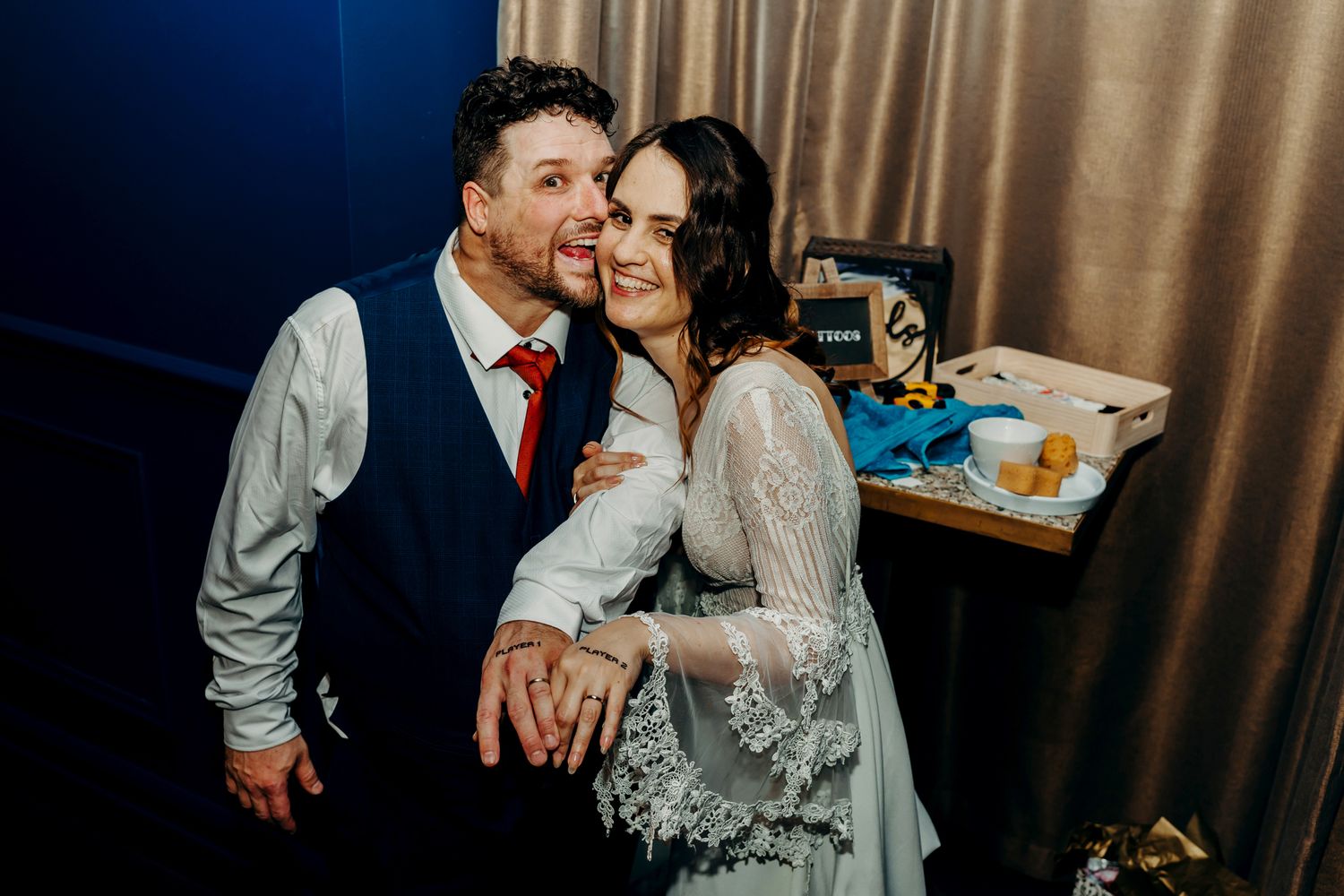 A couple smiling and embracing during their wedding celebration with reception decor in the background.