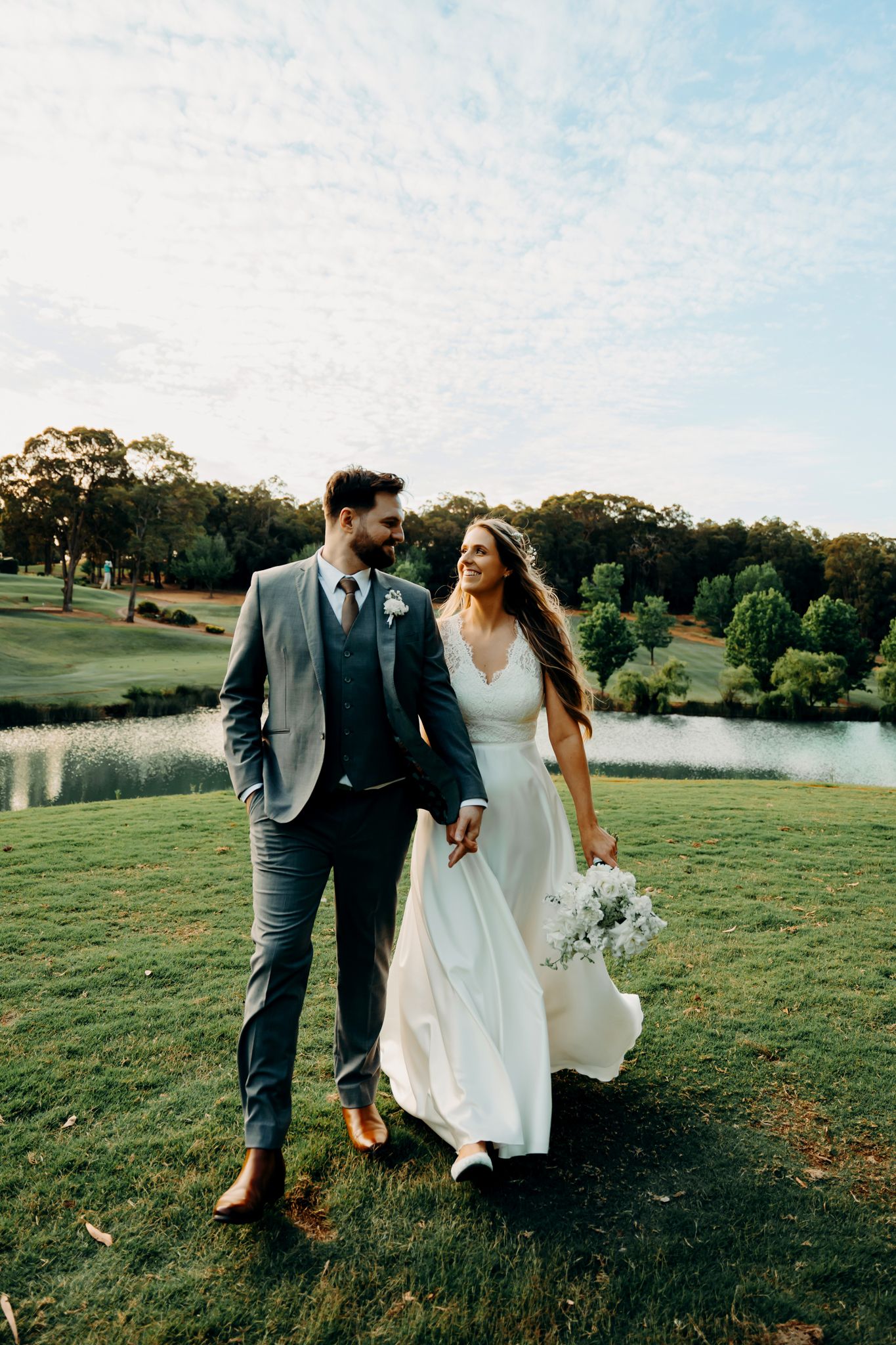 A couple walks together on a grassy hillside overlooking a lake during their outdoor wedding celebration.