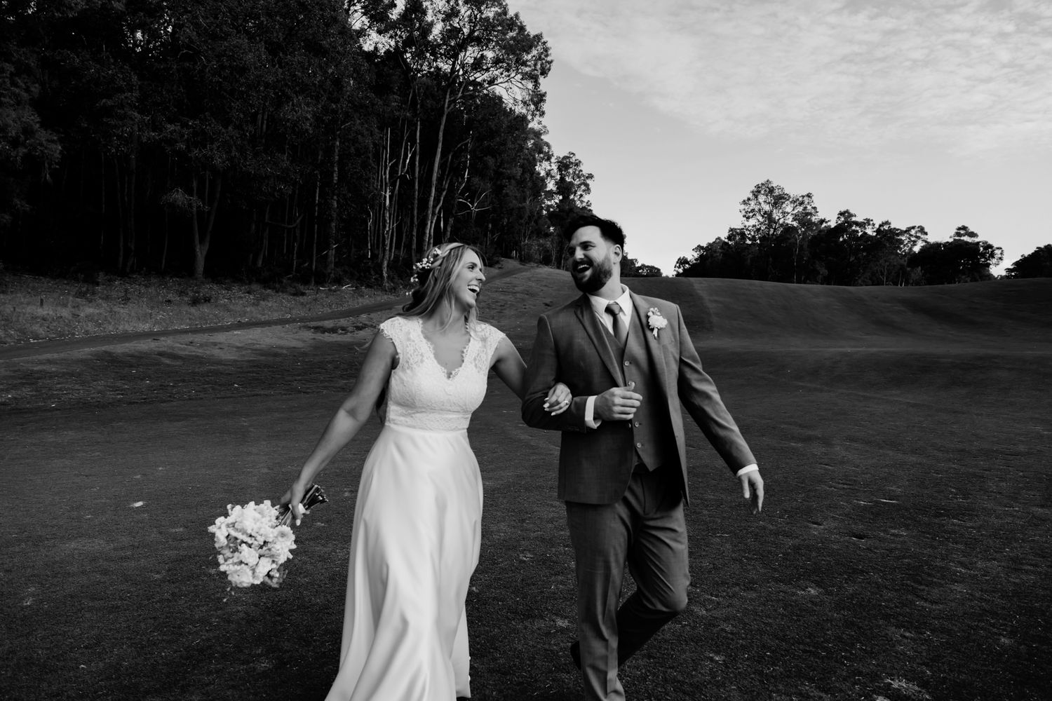 A couple walks together and laughs on a grassy hill with trees in the background during a wedding celebration.