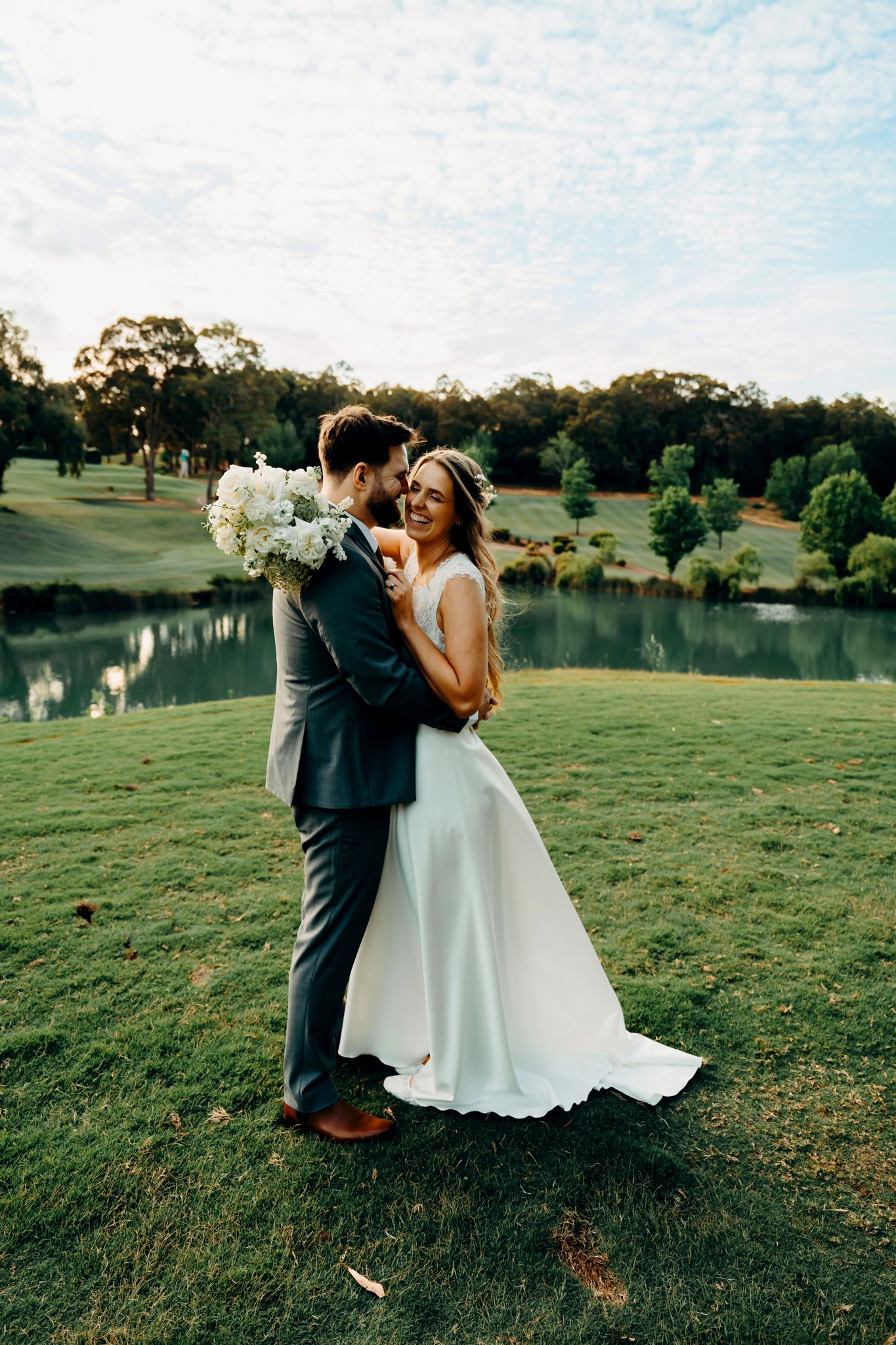 A romantic wedding photoshoot in a grassy field with a lake and trees in the background at sunset.