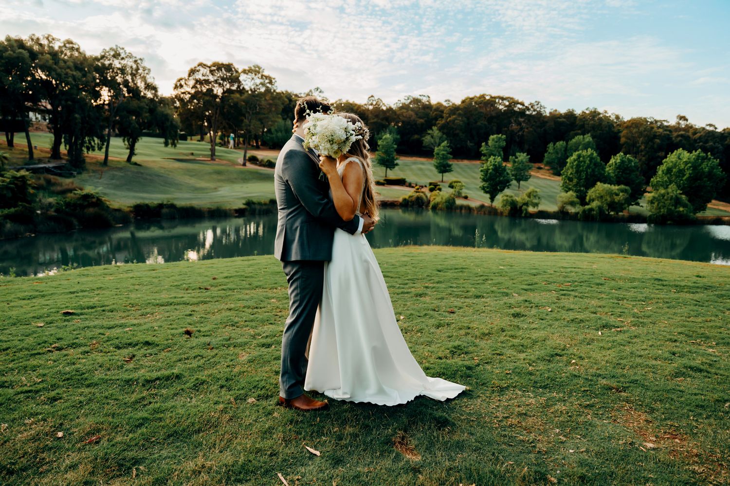A couple in wedding attire stands together on a lush green hillside overlooking a scenic lake at sunset.