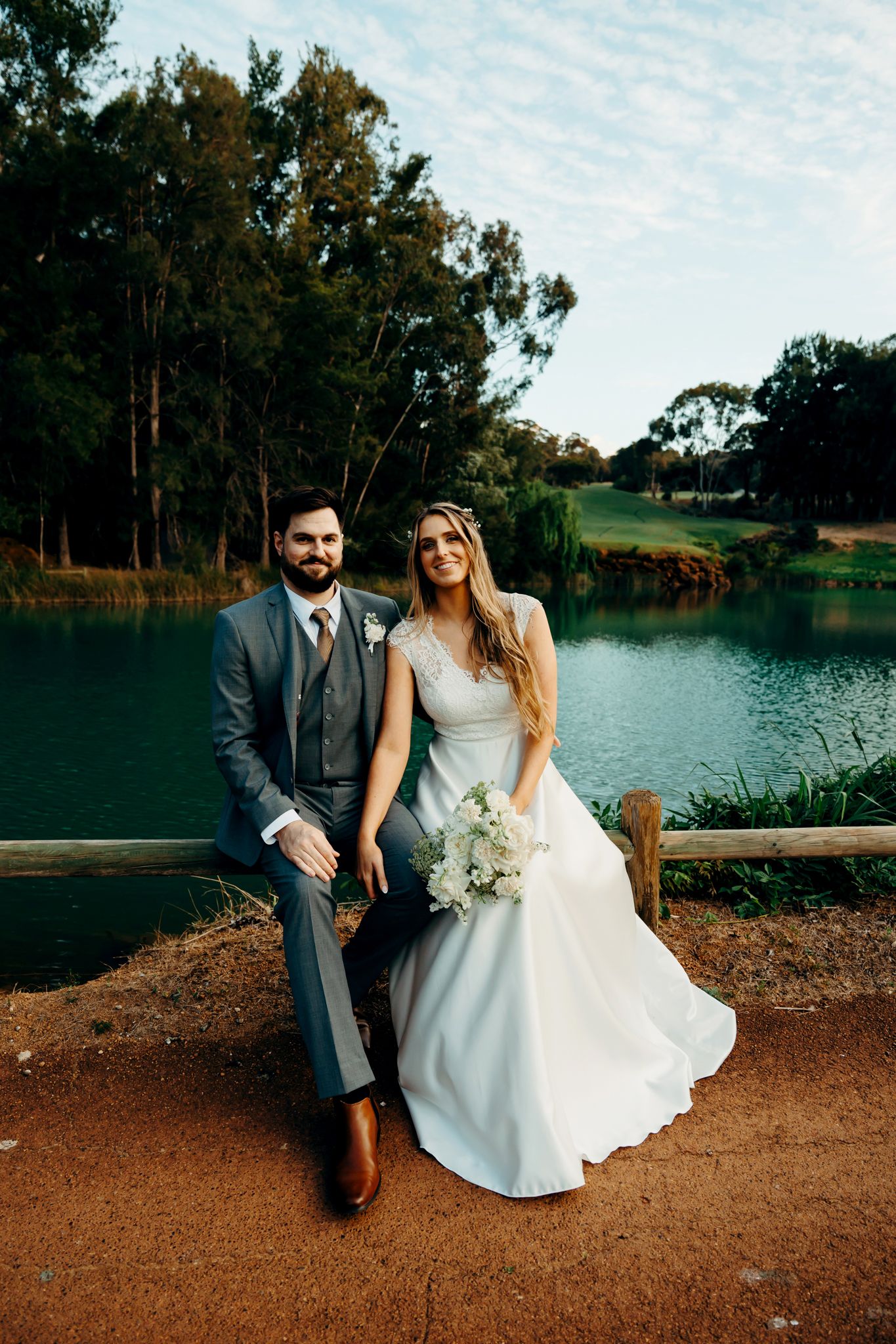 A wedding couple sits together on a dirt path by a serene lake surrounded by trees.