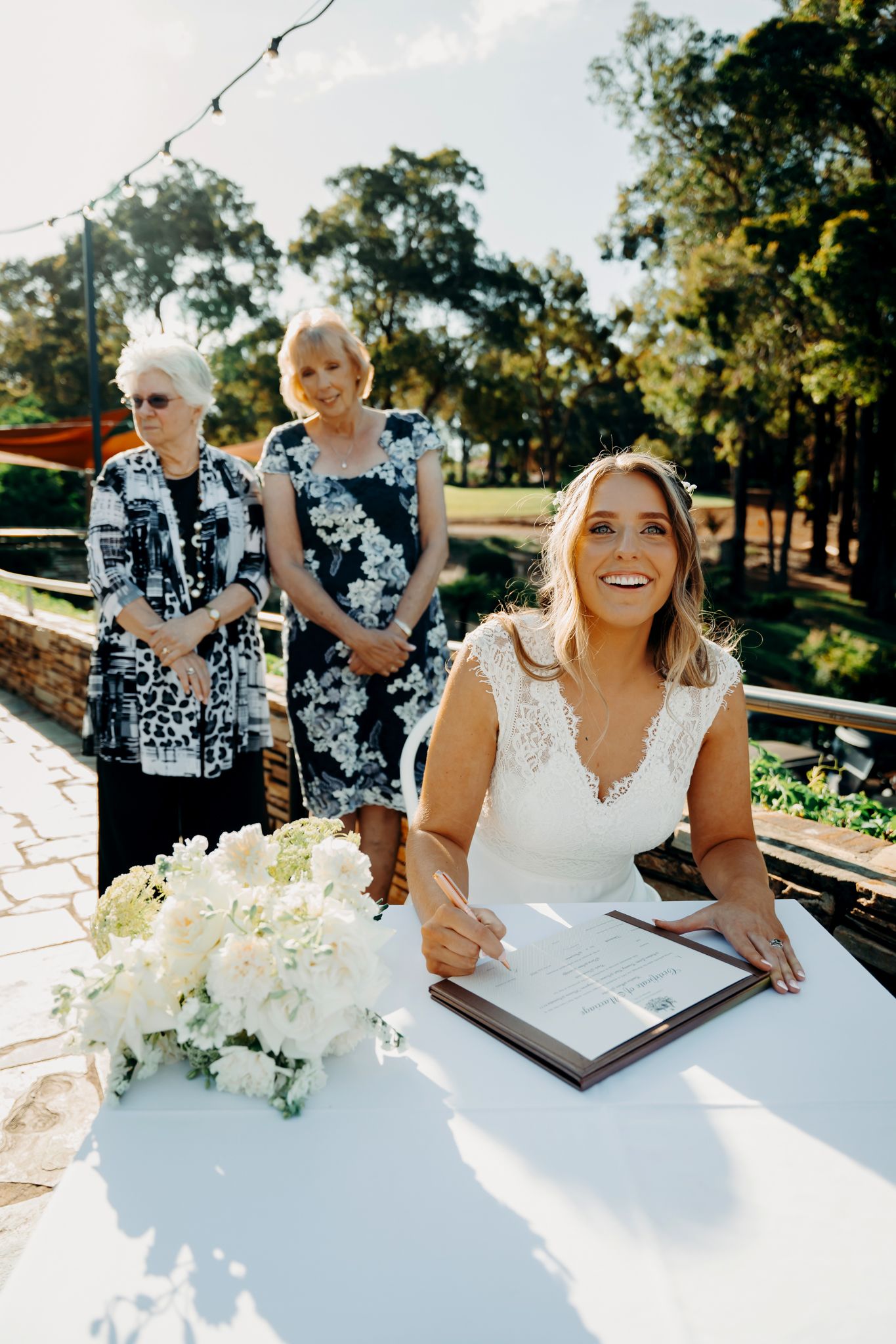 Wedding guests sign a white guestbook on an outdoor table with white floral decorations in the sunlight.