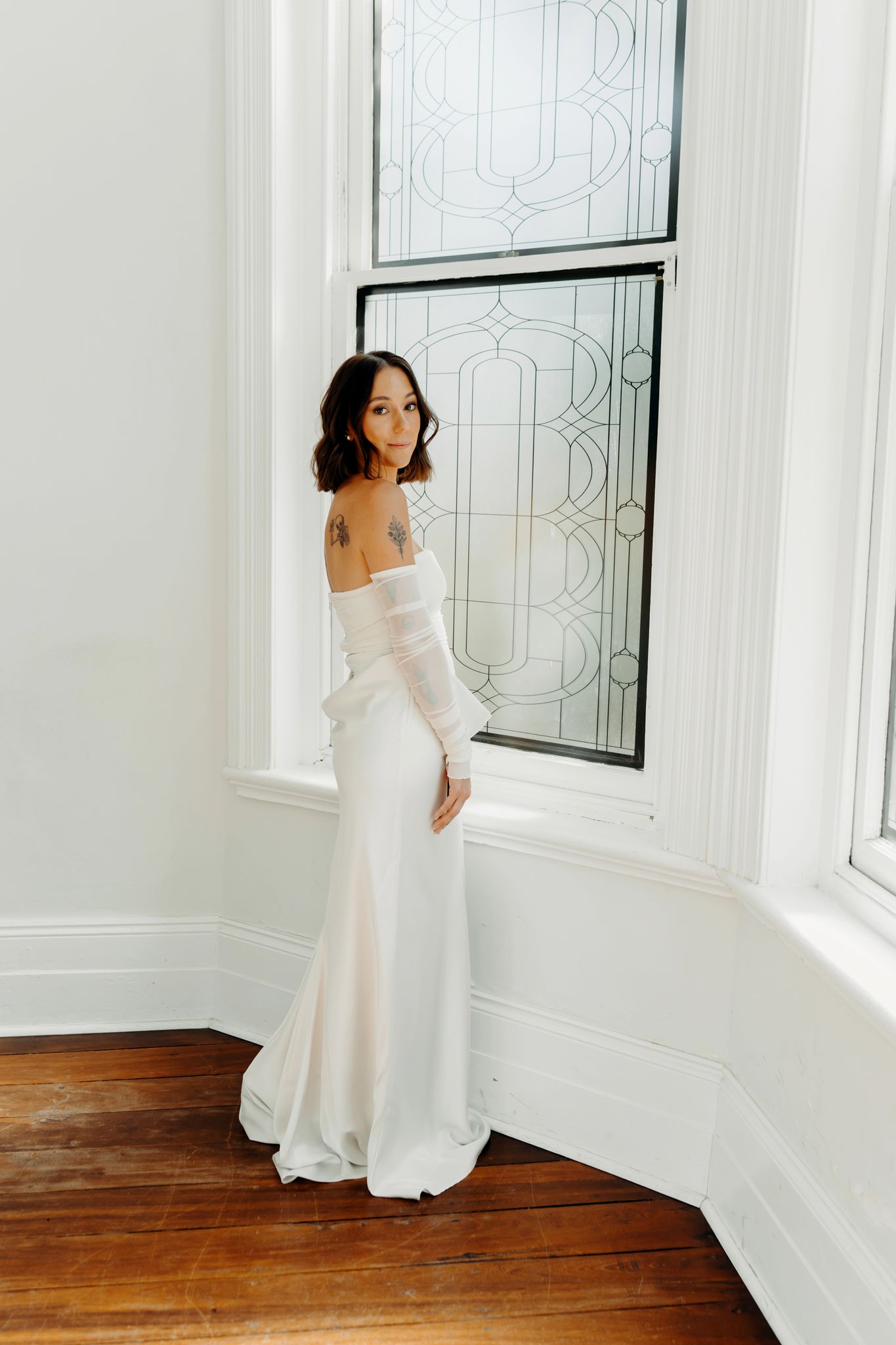 Woman in off-shoulder white wedding gown with long sleeves stands by ornate stained glass windows on hardwood floors.