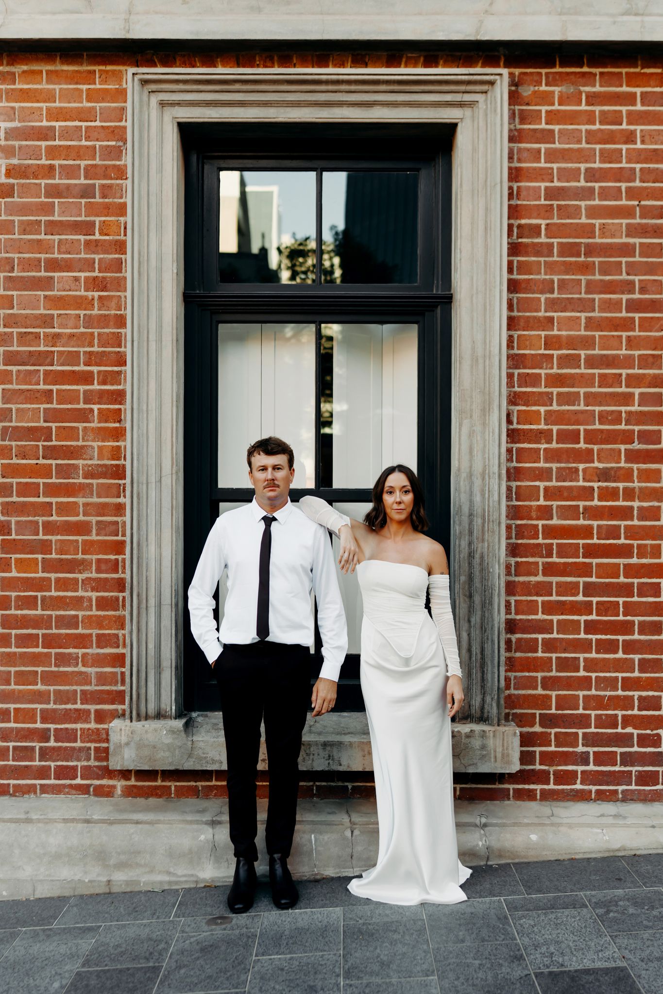 Bride in white off-shoulder gown and groom in black trousers and white shirt pose before a red brick wall.