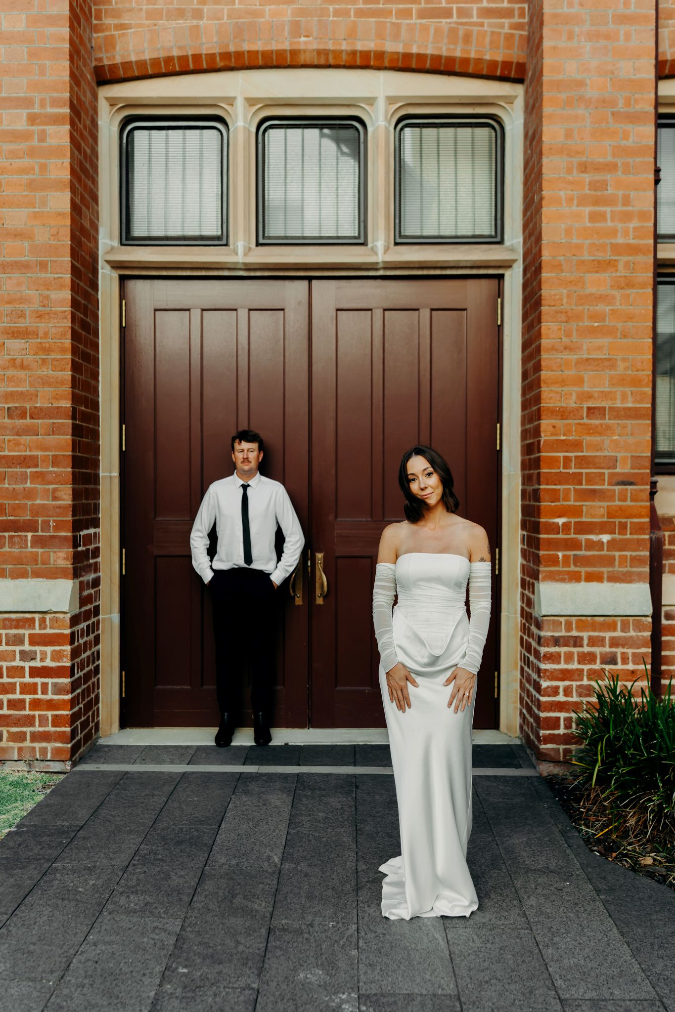 Bride in white off-shoulder gown stands before ornate brick building with groom in black tie suit leaning against doors.