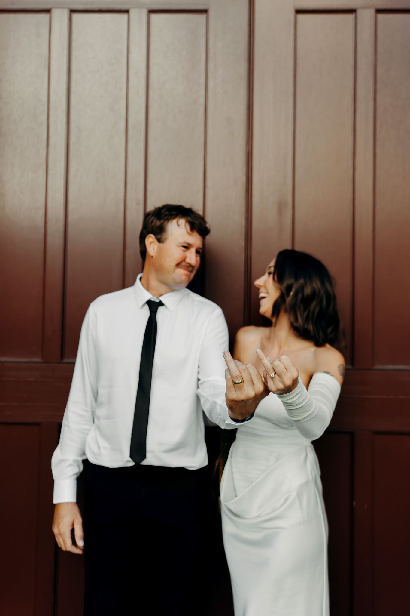 Newlywed couple laughing together in wedding attire, showing off rings, standing against a dark red wooden door.