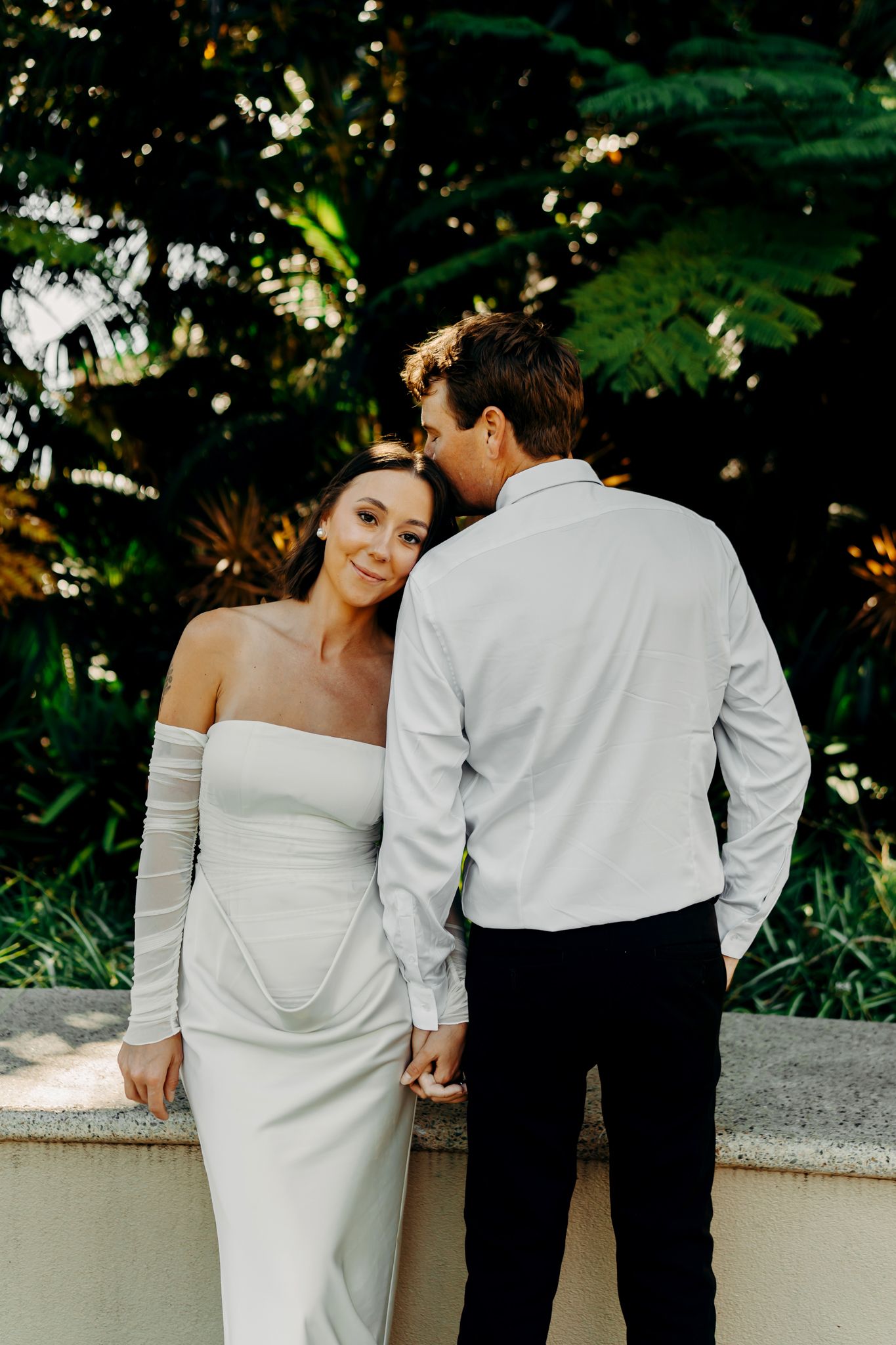 Bride in off-shoulder white gown leans on groom in white shirt, sharing tender moment outdoors.