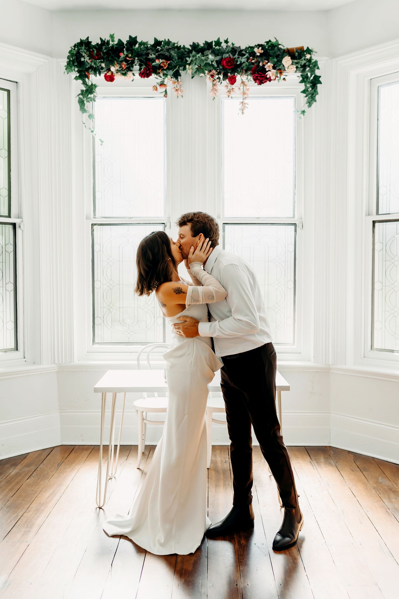 Couple sharing a romantic kiss in a bright white room with holiday garland above large windows on hardwood floors.