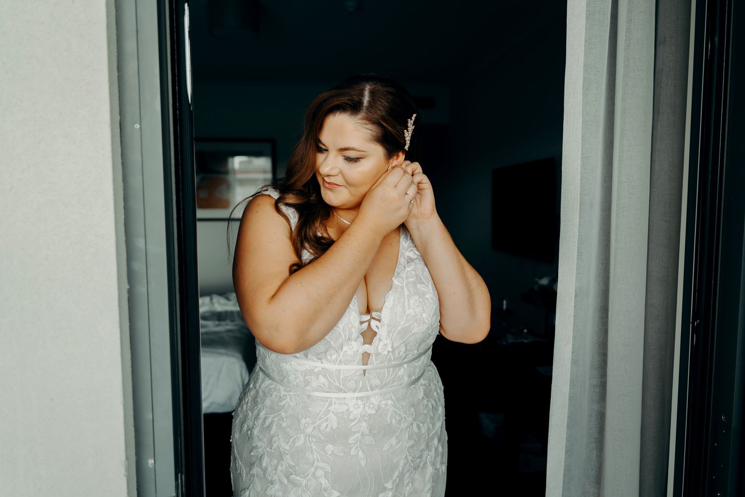 A bride in a white lace wedding dress adjusts her gown while getting ready in an elegant dressing room.